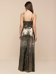 Wearing the Sayla Sequin Gown—a sleeveless, floor-length silk dress embellished with shimmering gold and olive green sequins—a woman with long brown hair stands with her back to the camera against a plain white backdrop, exuding vintage glamour.