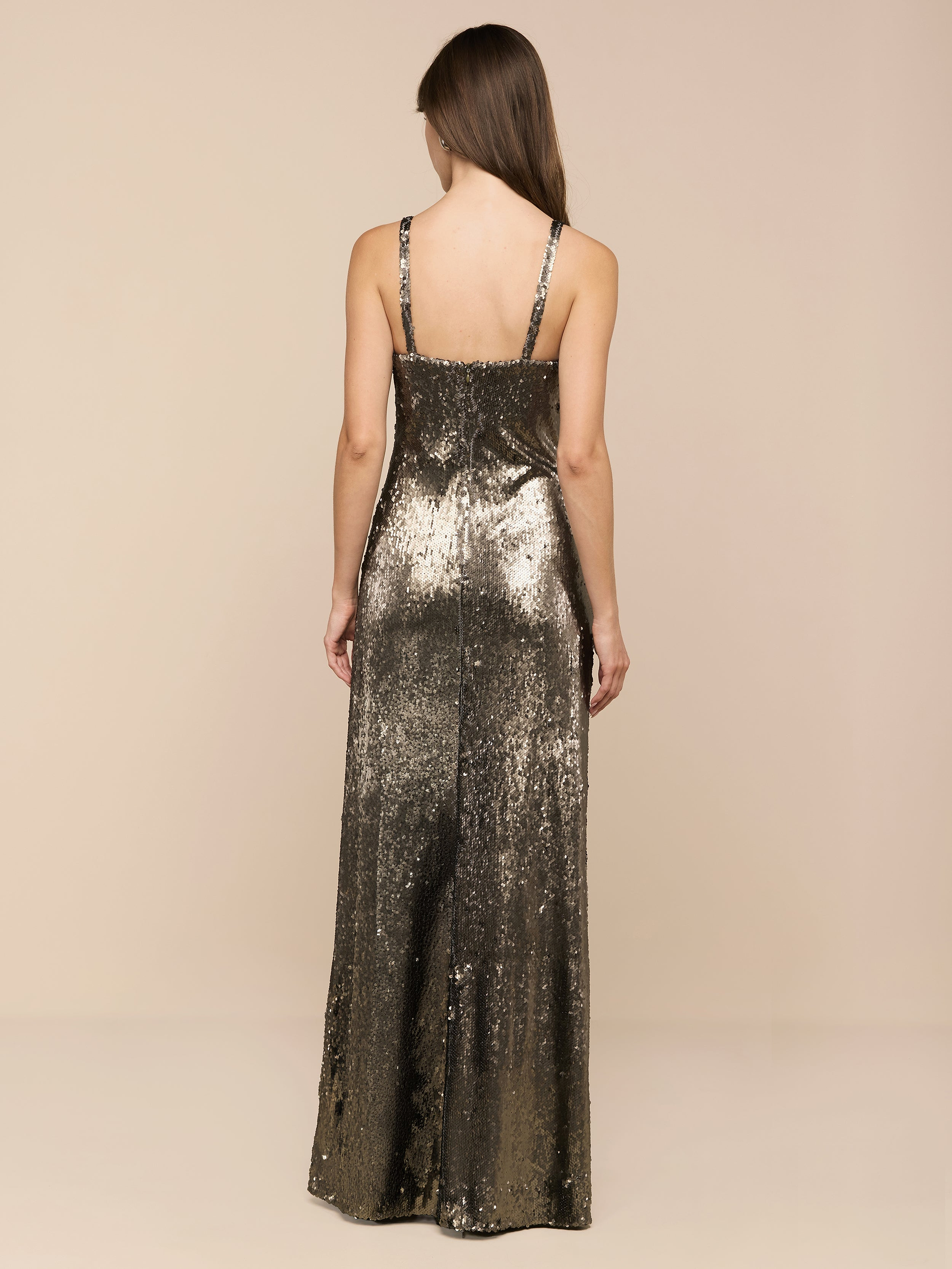 Wearing the Sayla Sequin Gown—a sleeveless, floor-length silk dress embellished with shimmering gold and olive green sequins—a woman with long brown hair stands with her back to the camera against a plain white backdrop, exuding vintage glamour.
