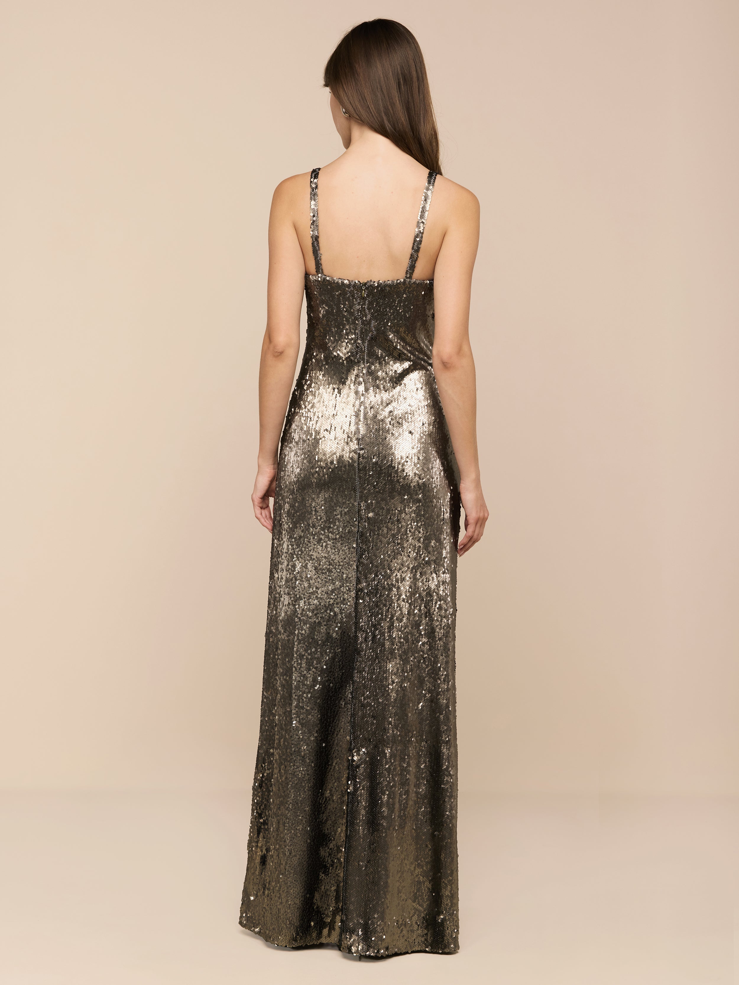 Wearing the Sayla Sequin Gown—a sleeveless, floor-length silk dress embellished with shimmering gold and olive green sequins—a woman with long brown hair stands with her back to the camera against a plain white backdrop, exuding vintage glamour.