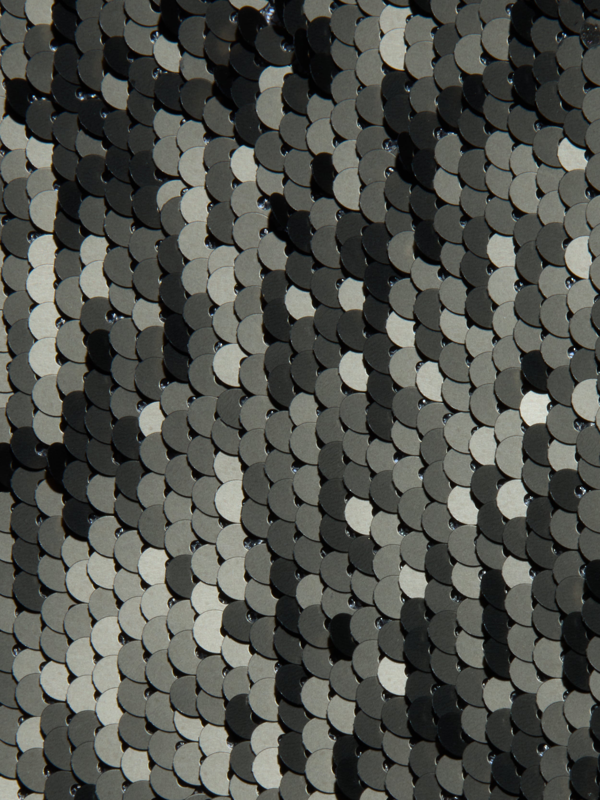 Close-up of overlapping black, silver, and olive green sequins on the Sayla Sequin Gown creates a shiny, textured pattern with a wavy, reflective effect reminiscent of vintage glamour.