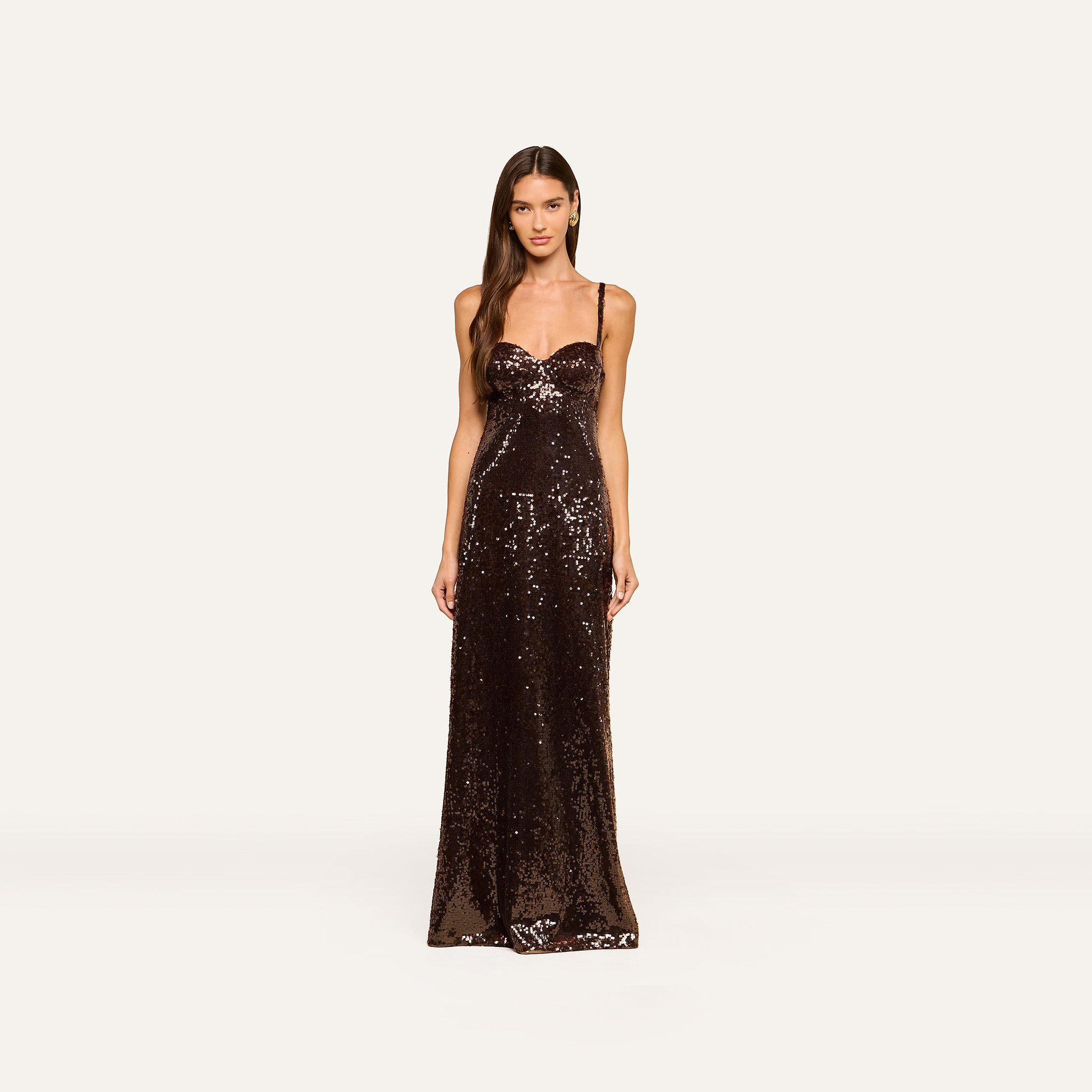 A woman stands facing forward, wearing the Sayla Sequin Gown—a floor-length, event-ready dress featuring thin straps and brown sequins—against a plain, light background.