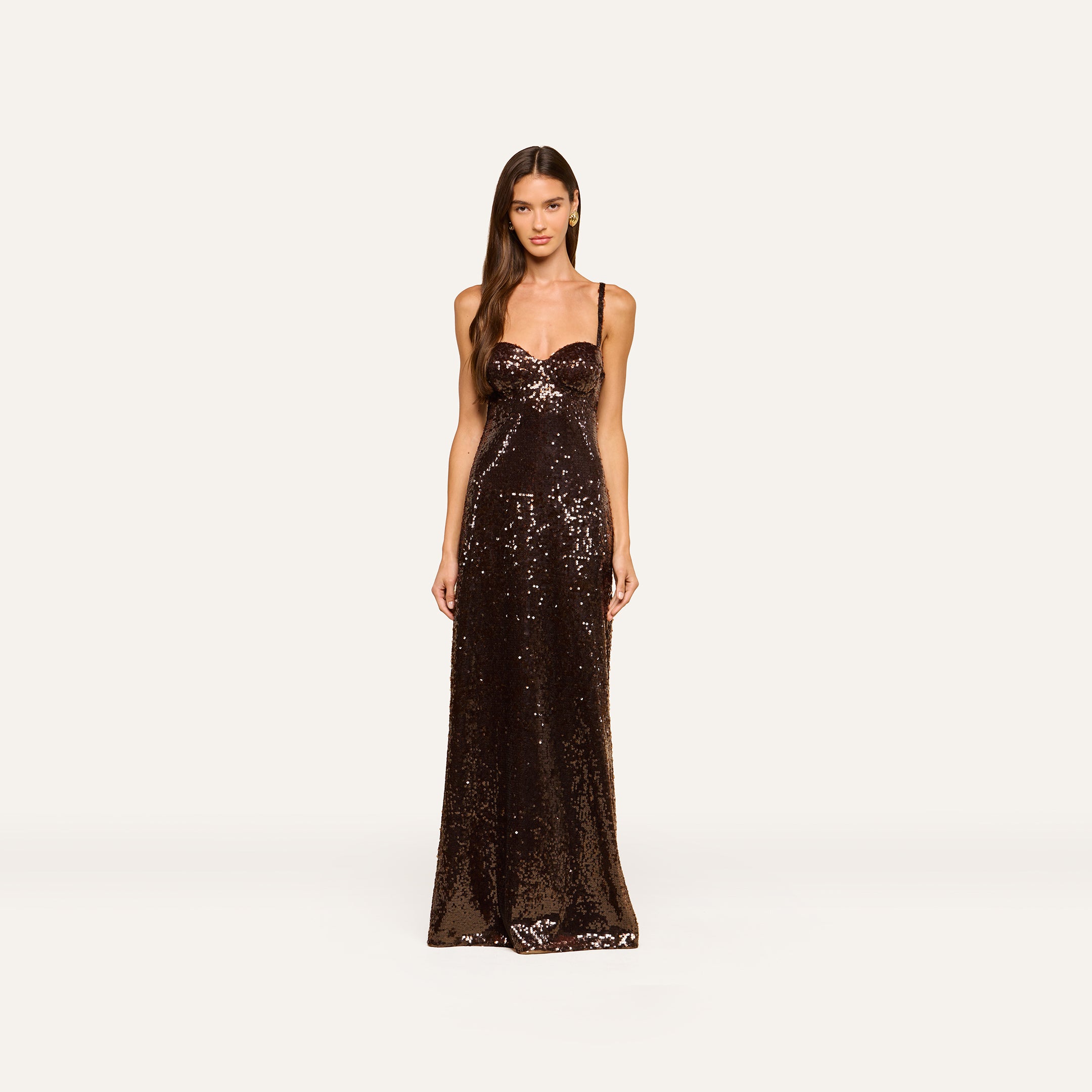 A woman stands facing forward, wearing the Sayla Sequin Gown—a floor-length, event-ready dress featuring thin straps and brown sequins—against a plain, light background.