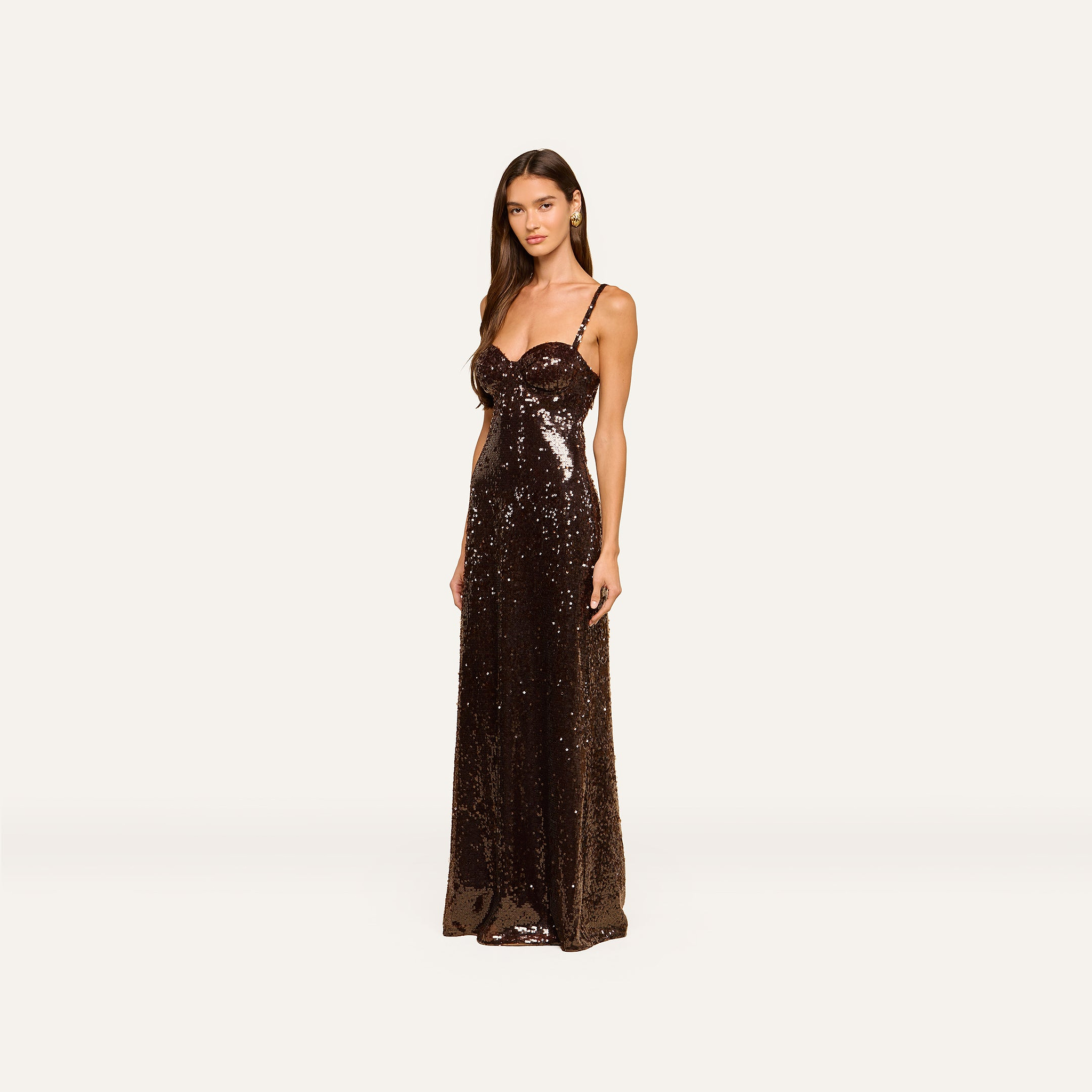 A woman stands against a plain, light background wearing the Sayla Sequin Gown—a dark brown, floor-length dress with thin straps. This event-ready gown exudes timeless elegance.