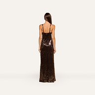 A woman with long straight hair stands facing away, wearing the Sayla Sequin Gown—a floor-length dress in dark brown sequins with thin shoulder straps—against a plain light background, creating an event-ready look perfect for stylish occasions.
