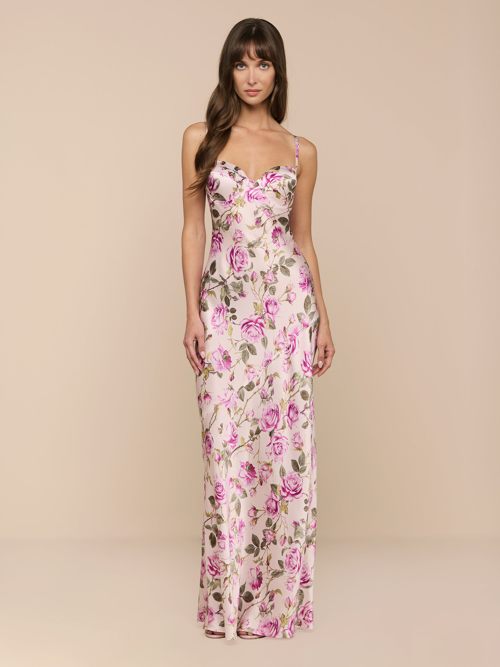 Una donna dai lunghi capelli castani indossa labito Sayla Silk Gown — un abito lungo senza maniche, rosa a fiori, con spalline sottili — che evoca il fascino vintage mentre posa su uno sfondo beige a tinta unita, guardando direttamente nella fotocamera.