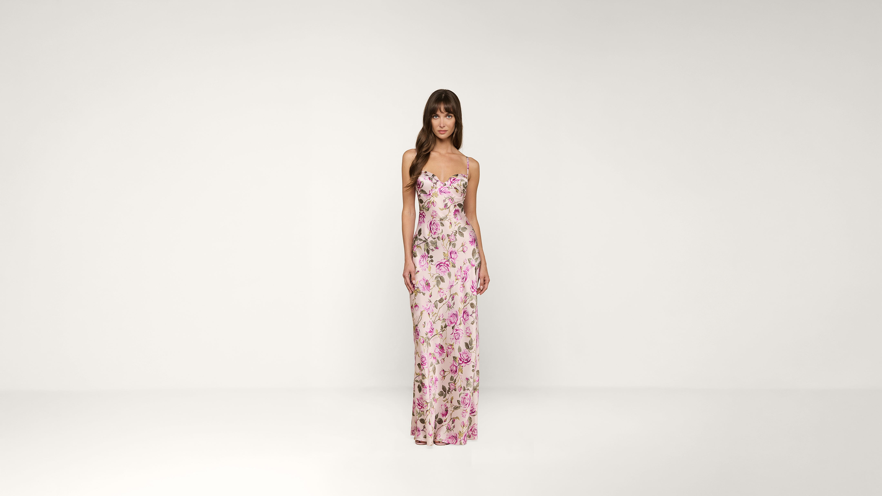 A woman stands against a plain white background, wearing a long, fitted pink floral dress with thin straps.