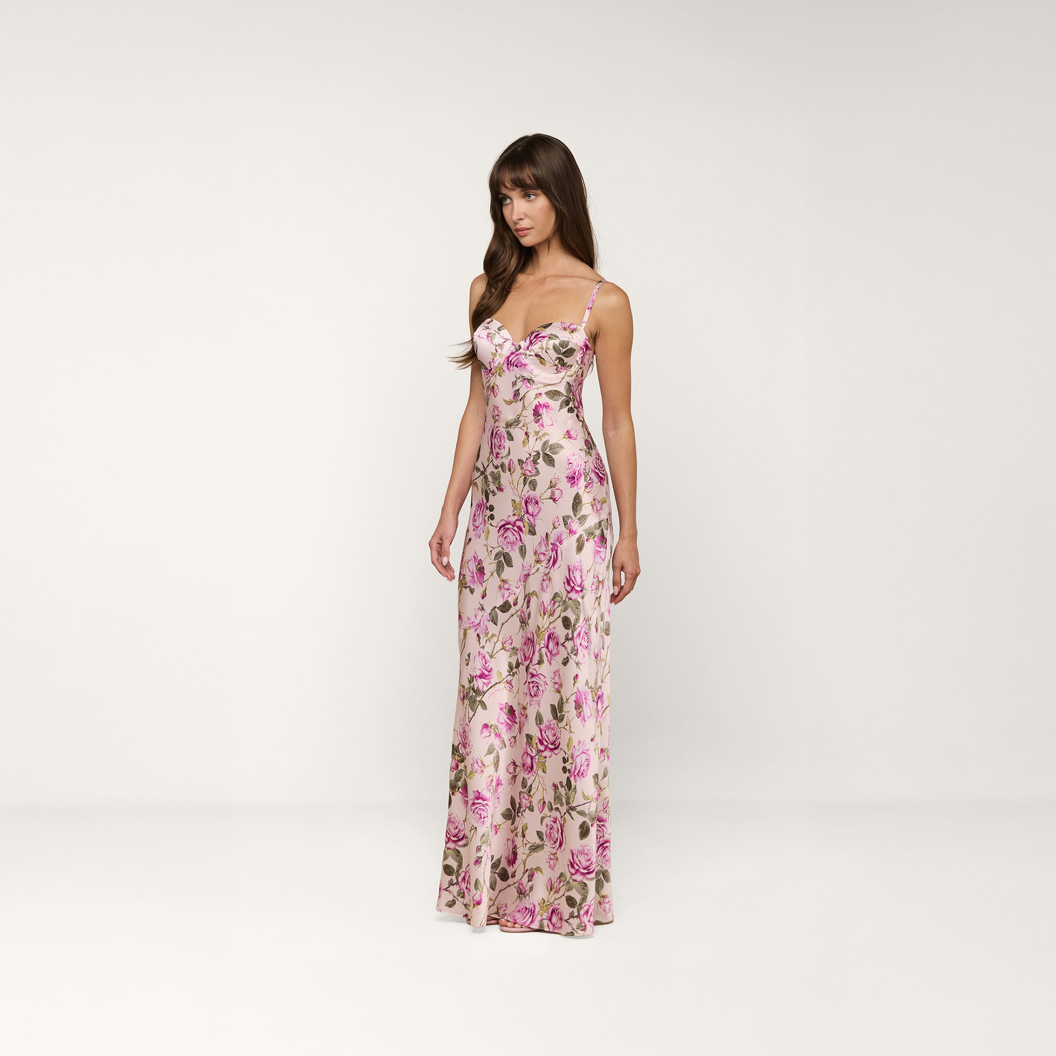 Una donna indossa labito Sayla Silk Gown, un abito lungo senza maniche con spalline sottili e una stampa di rose rosa e viola, in piedi su uno sfondo beige a tinta unita che evoca il fascino vintage.