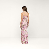 A woman stands with her back to the camera, wearing the Sayla Silk Gown, which features a long silhouette adorned with a purple rose print and pink flowers on a light background, set against a plain white backdrop.