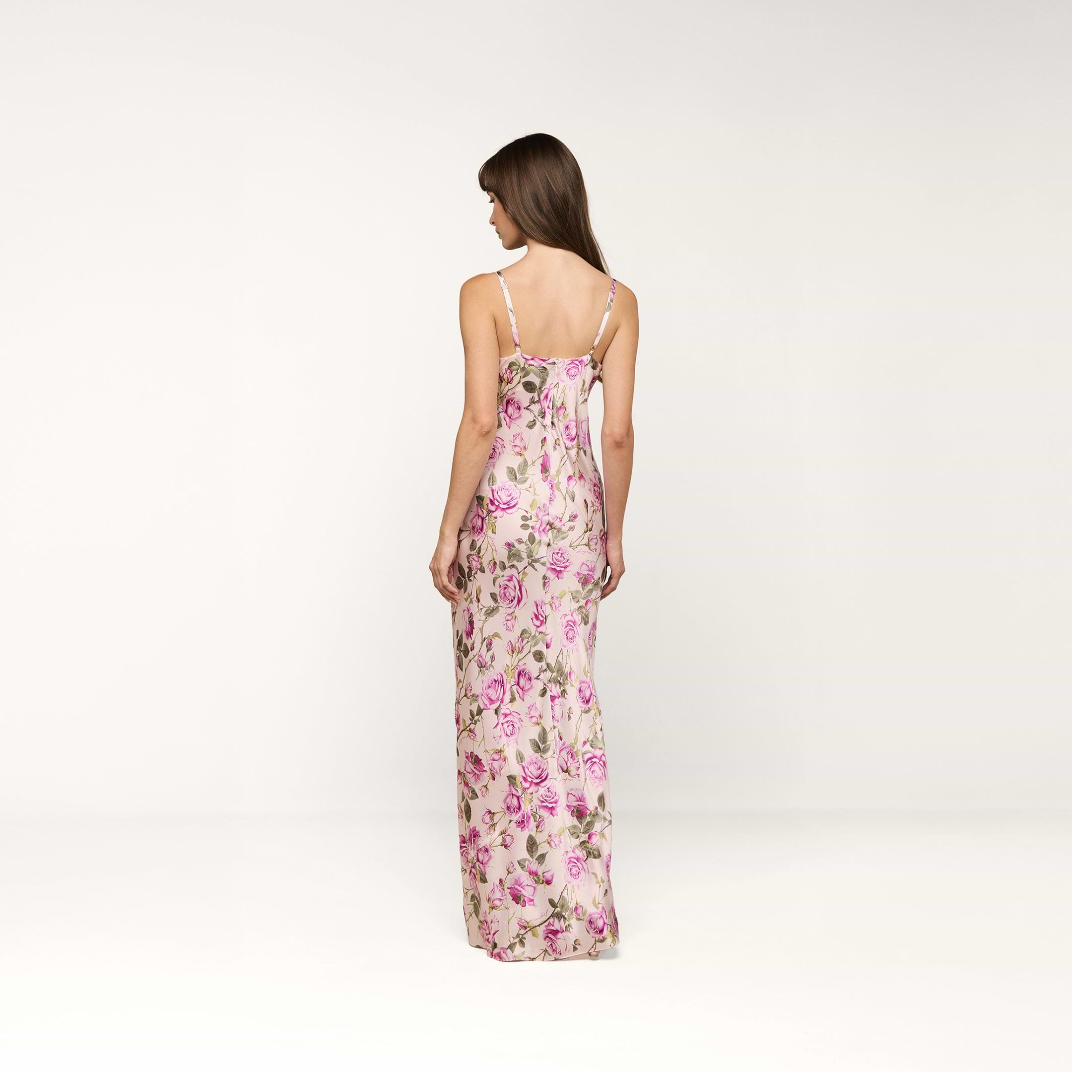 A woman stands with her back to the camera, wearing the Sayla Silk Gown, which features a long silhouette adorned with a purple rose print and pink flowers on a light background, set against a plain white backdrop.