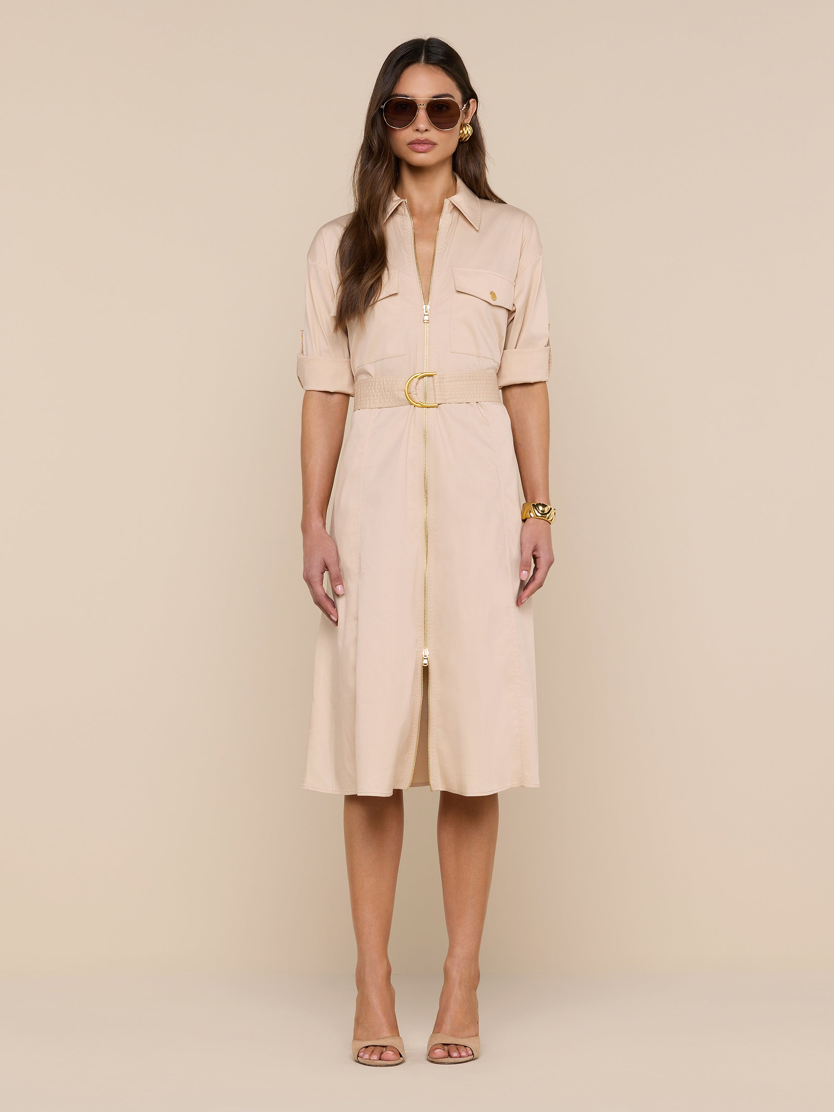 A woman wearing sunglasses and the Callen Shirt Dress—a light beige, belted design—stands against a plain white background, styled with neutral high-heeled sandals and gold bracelets.