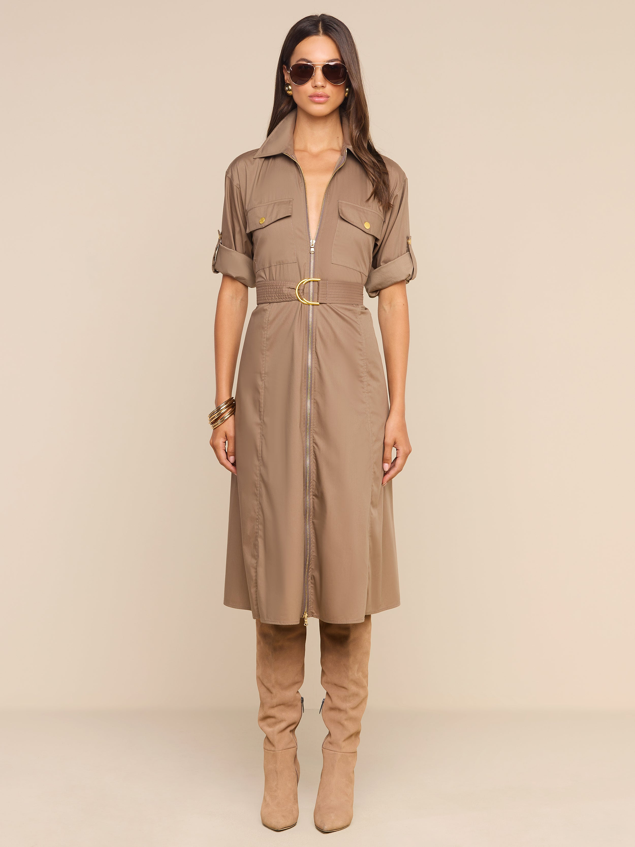 A woman stands against a plain white background, hands by her sides, wearing the Callen Shirt Dress—a tan cotton-blend poplin style with an exposed front zip—paired with knee-high tan boots and sunglasses.