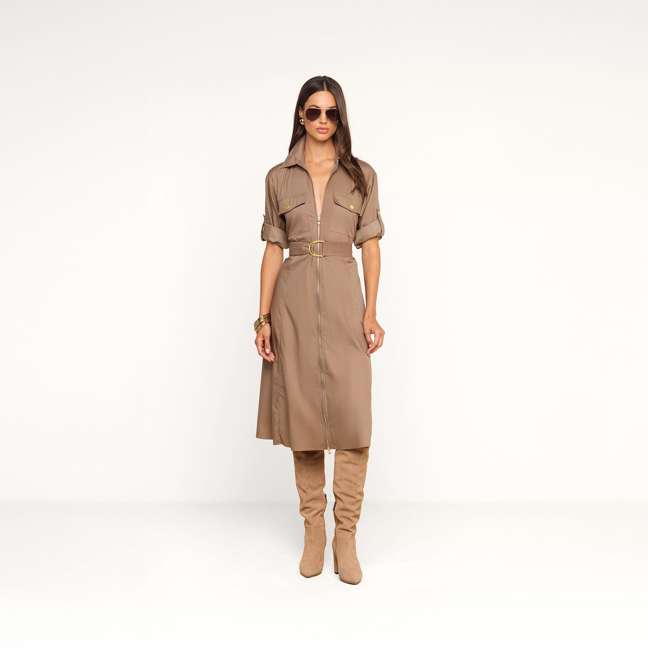A woman wears the Callen Shirt Dress—a tan belted shirt dress with rolled sleeves and a front zip detail—styled with tan knee-high boots, sunglasses, and bracelets, standing against a plain white background.