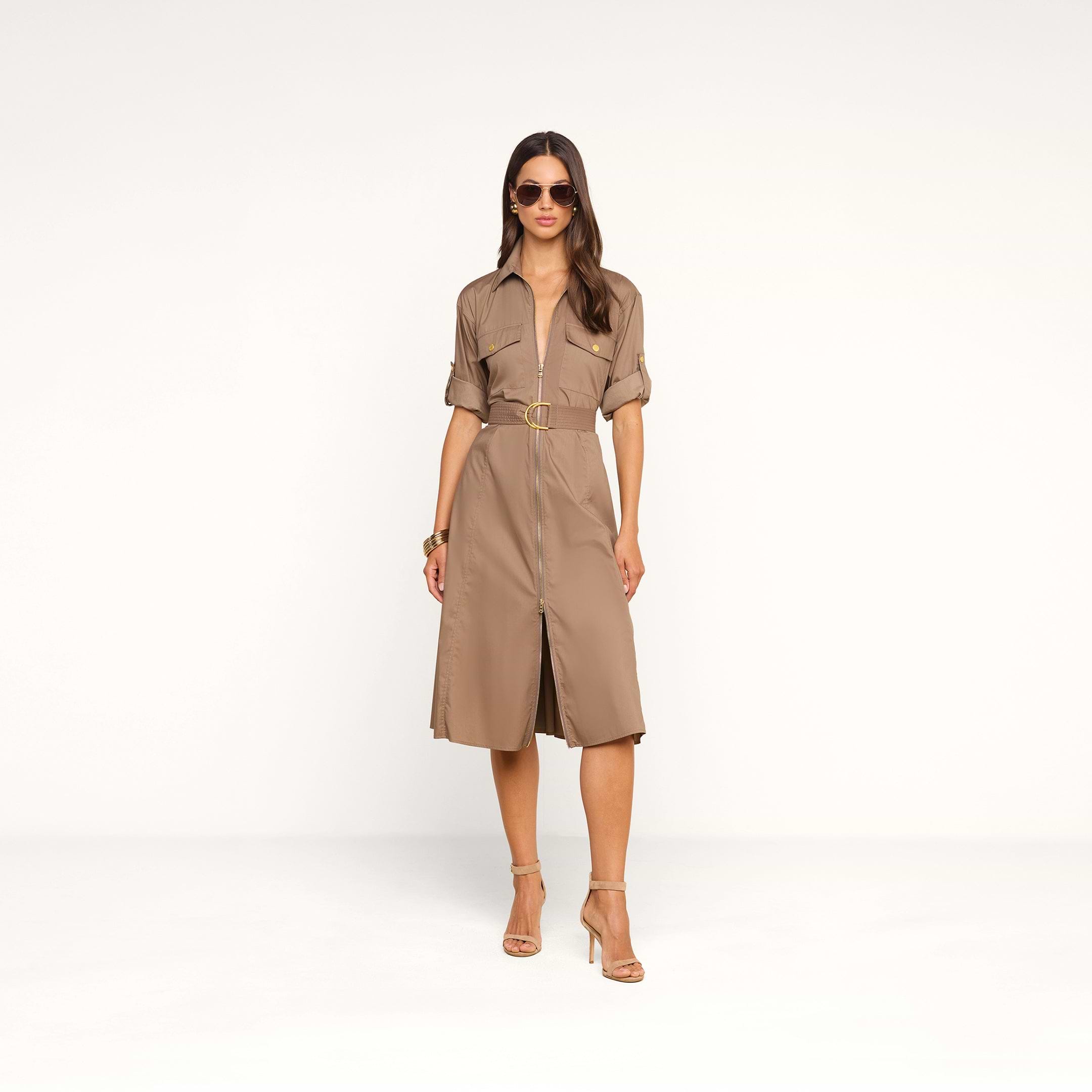 A woman wearing sunglasses and high-heeled sandals stands confidently in a studio against a plain white background, modeling the Callen Shirt Dress—a tan cotton-blend poplin shirt dress featuring an exposed front zip and rolled sleeves.