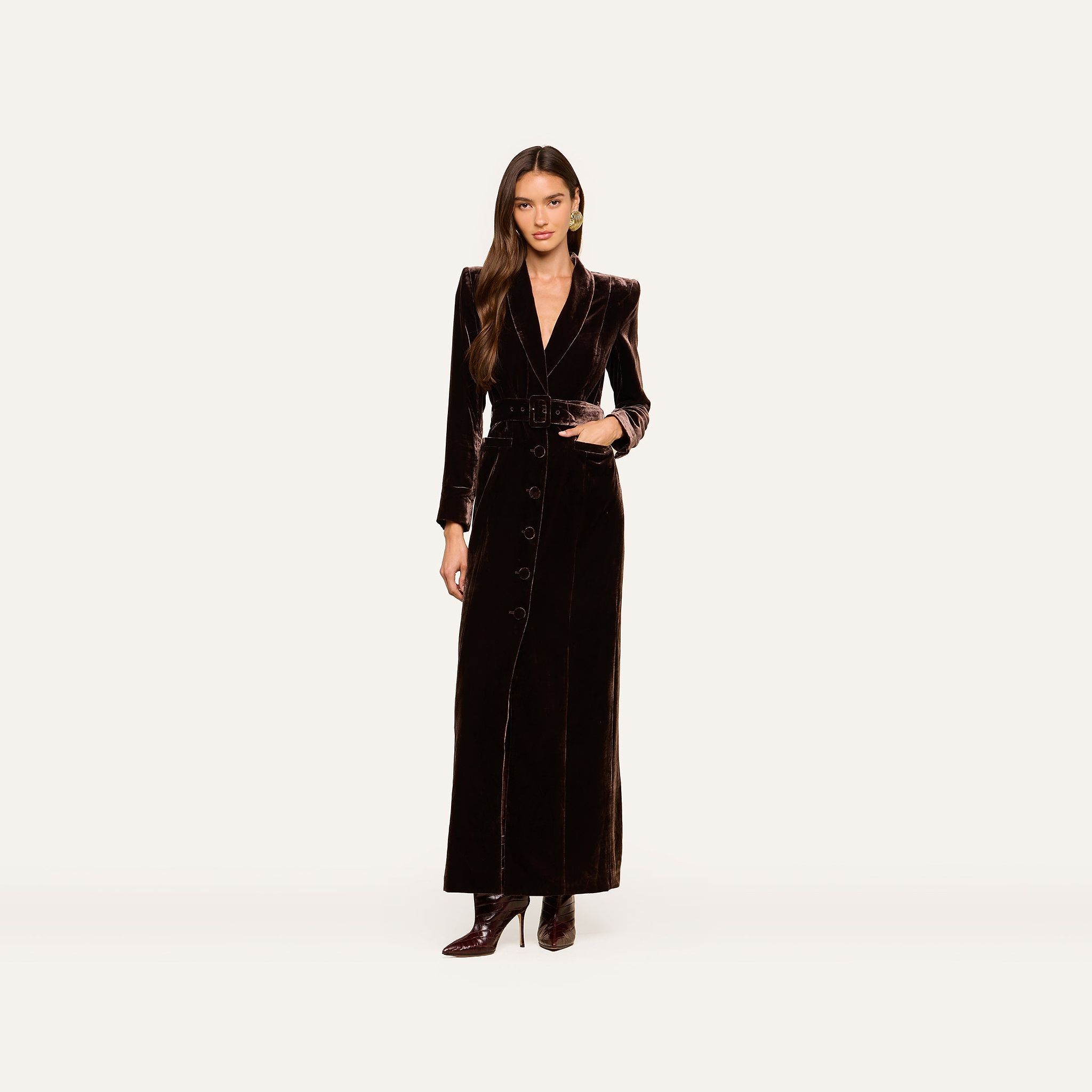 A woman wearing the Raini Velvet Maxi Dress—a long black dress with buttons, a belted waist, and a shawl collar—stands against a plain white background pairing it with high-heeled boots for a chic Fall 2026-inspired look.