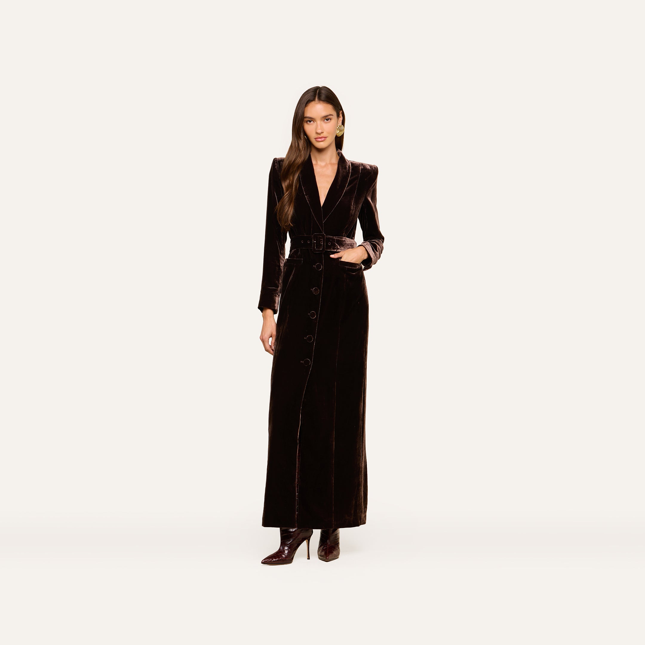 A woman wearing the Raini Velvet Maxi Dress—a long black dress with buttons, a belted waist, and a shawl collar—stands against a plain white background pairing it with high-heeled boots for a chic Fall 2026-inspired look.