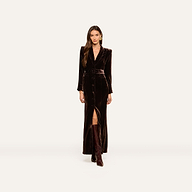 Wearing the Raini Velvet Maxi Dress with a matching belt and knee-high boots, a woman stands against a plain light background; the stylish velvet dress features a shawl collar.