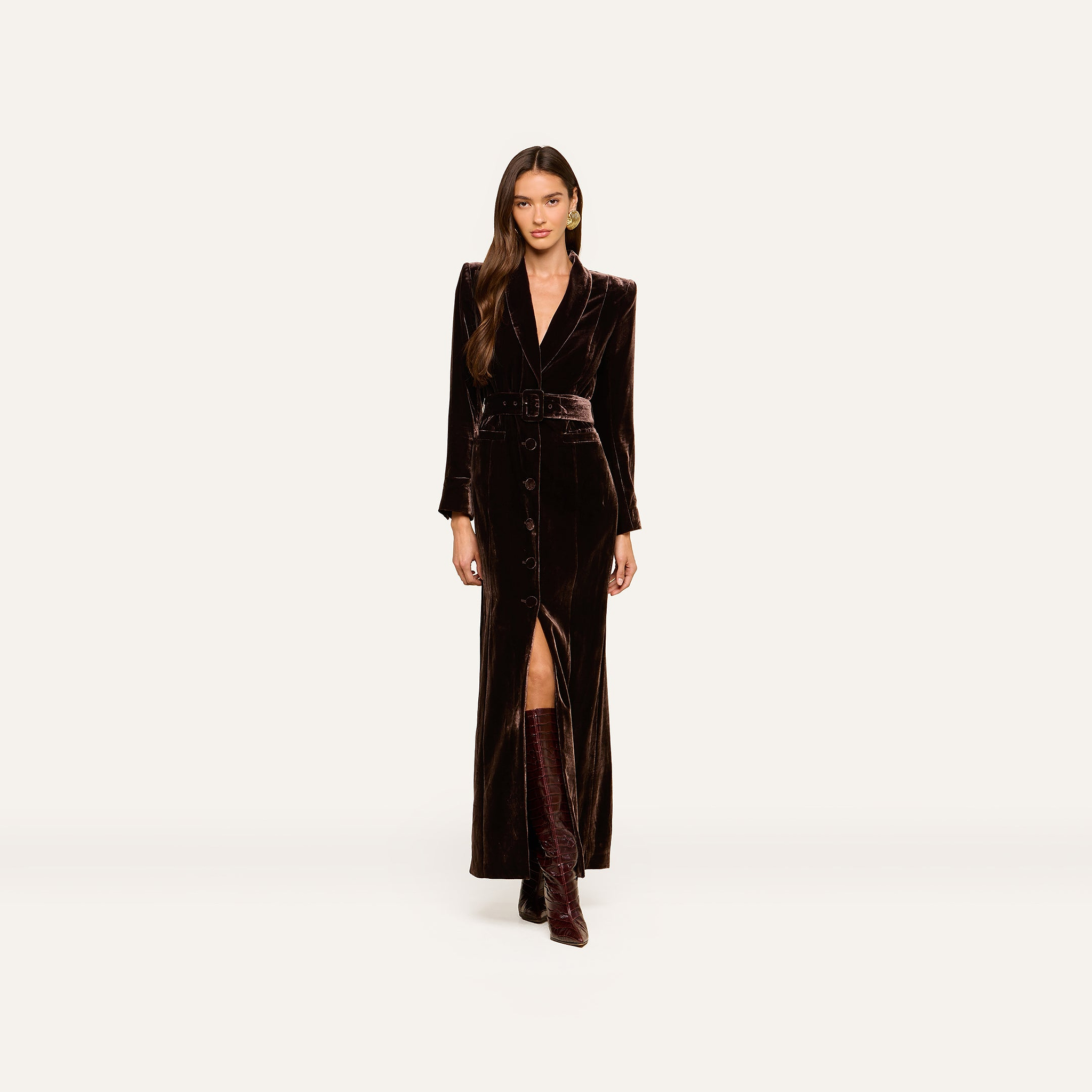 Wearing the Raini Velvet Maxi Dress with a matching belt and knee-high boots, a woman stands against a plain light background; the stylish velvet dress features a shawl collar.