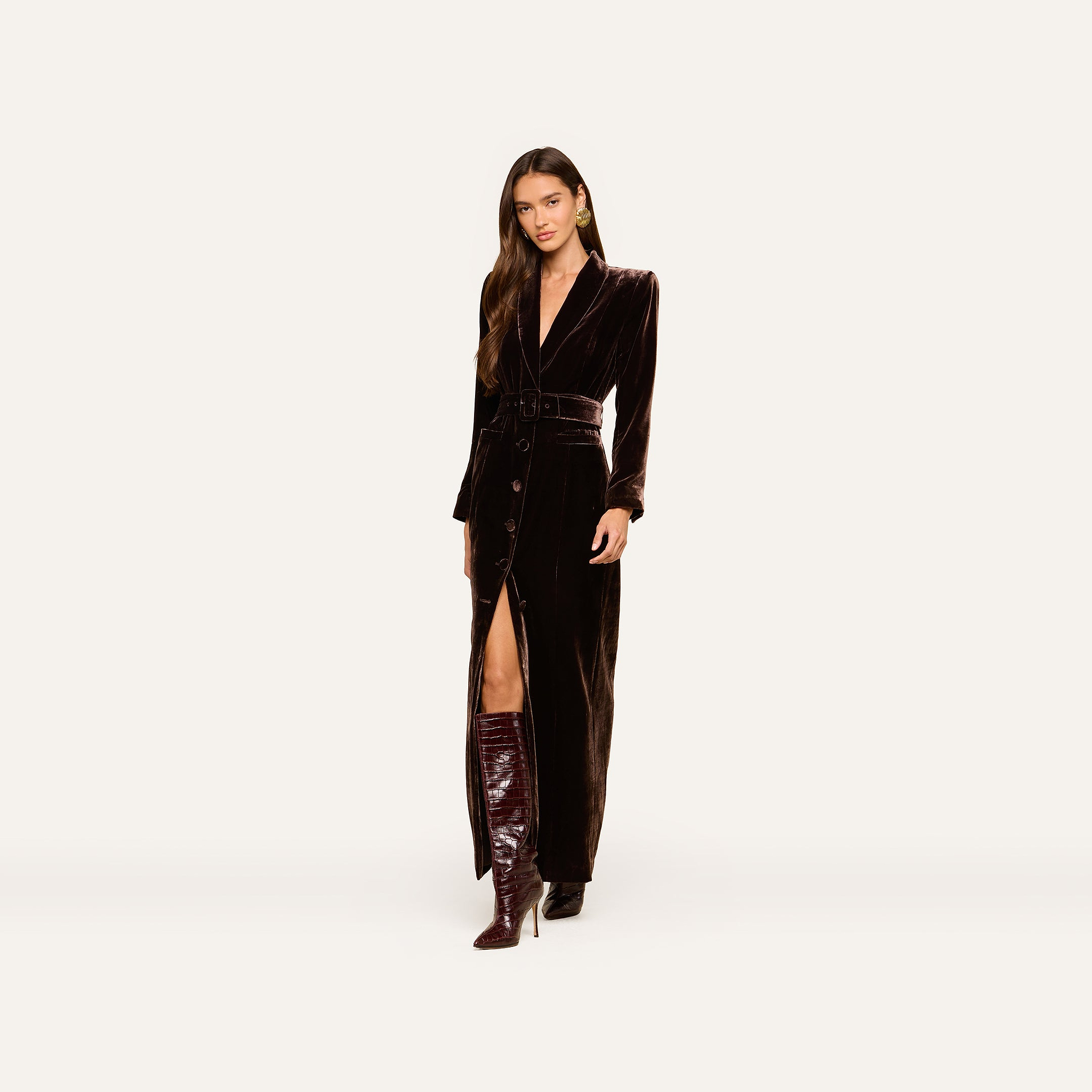 A woman stands facing forward in the Raini Velvet Maxi Dress, a long, dark gown featuring a shawl collar and high slit. She pairs it with tall, shiny brown boots and wears her long hair loose. The plain, light-colored background highlights the Fall 2026 fashion trend.