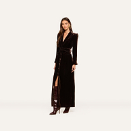 Wearing the Raini Velvet Maxi Dress—a long, dark silhouette with a shawl collar and belt—she pairs it with knee-high, pointed boots against a simple, light backdrop for a chic nod to Fall 2026 fashion.