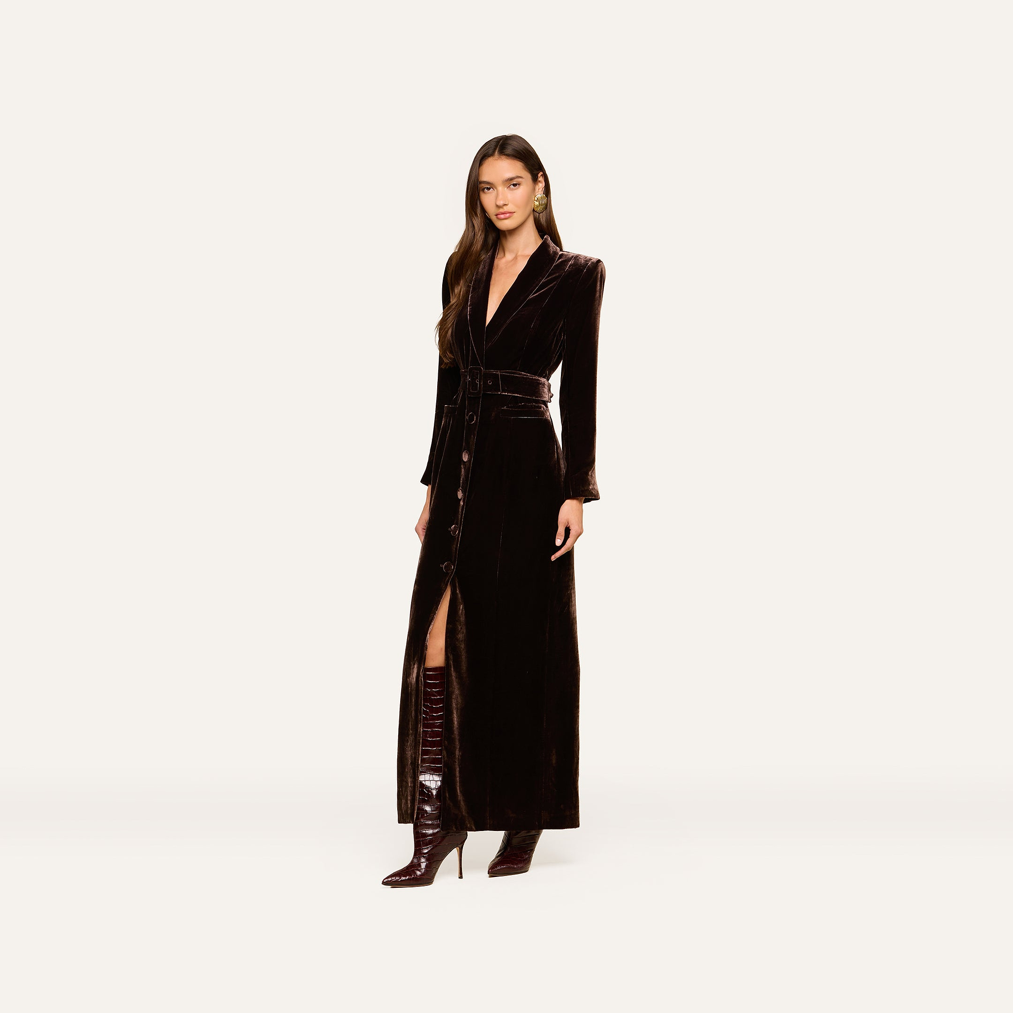Wearing the Raini Velvet Maxi Dress—a long, dark silhouette with a shawl collar and belt—she pairs it with knee-high, pointed boots against a simple, light backdrop for a chic nod to Fall 2026 fashion.