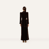 A woman stands facing away, dressed in the Raini Velvet Maxi Dress featuring a shawl collar, long sleeves, and a belted waist. She pairs it with dark heels and has her straight hair tied back. The plain, light-colored background highlights the elegance of Fall 2026 fashion.