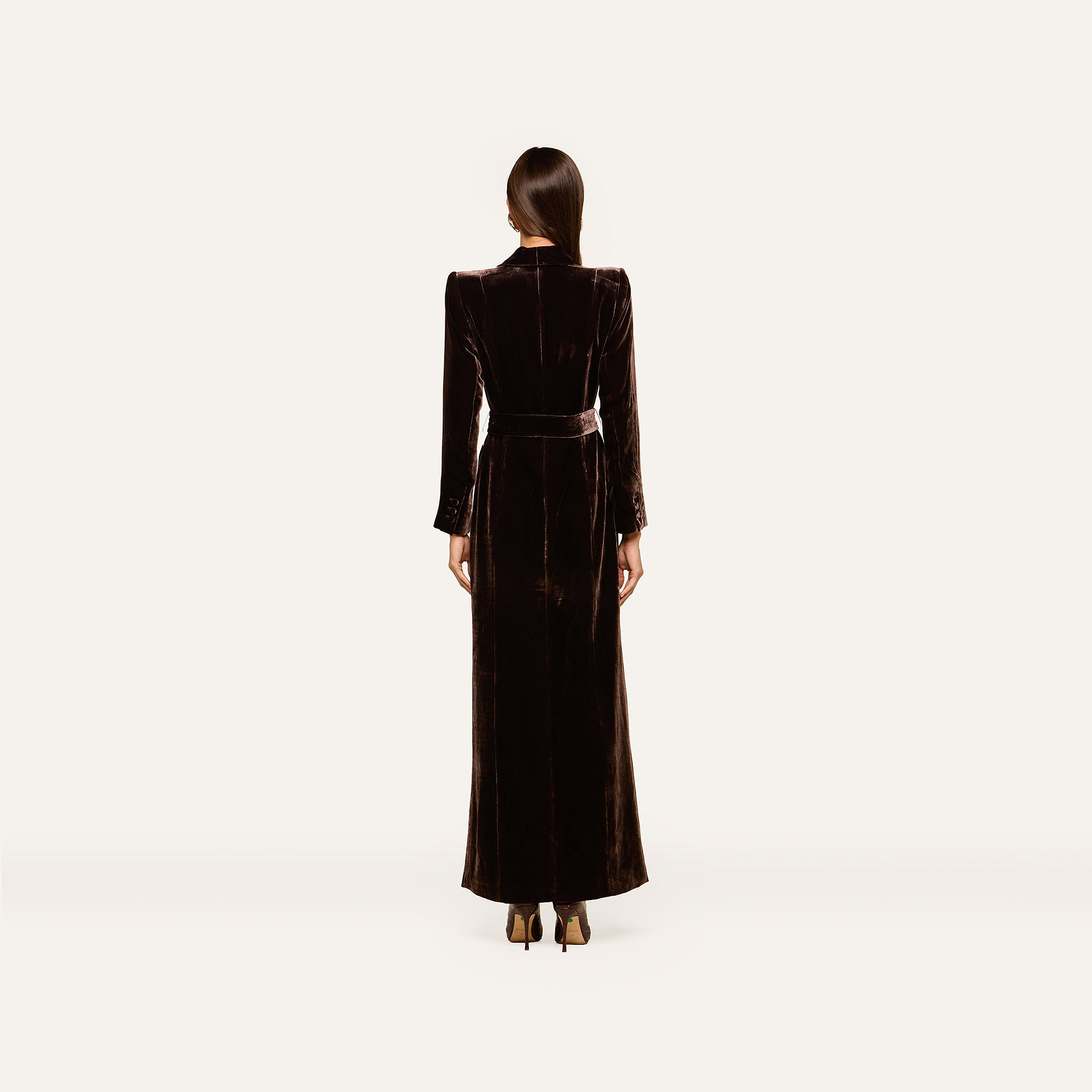 A woman stands facing away, dressed in the Raini Velvet Maxi Dress featuring a shawl collar, long sleeves, and a belted waist. She pairs it with dark heels and has her straight hair tied back. The plain, light-colored background highlights the elegance of Fall 2026 fashion.