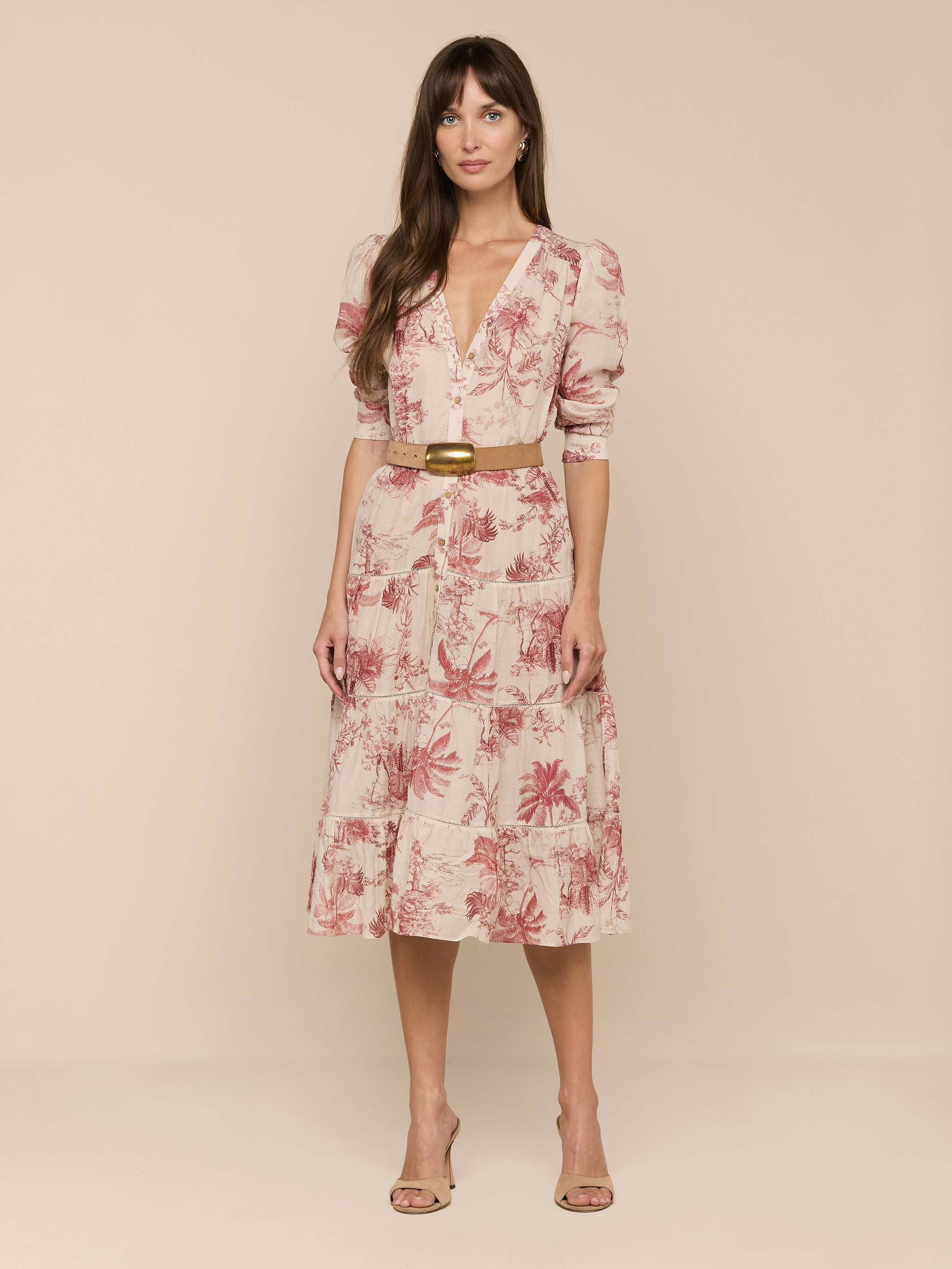 A woman with long brown hair stands against a plain white background wearing the Serafine Midi Dress, a light pink floral style featuring lightweight voile fabric, a V-neckline, puff sleeves, and a coordinating tan belt. She completes her look with nude heels.