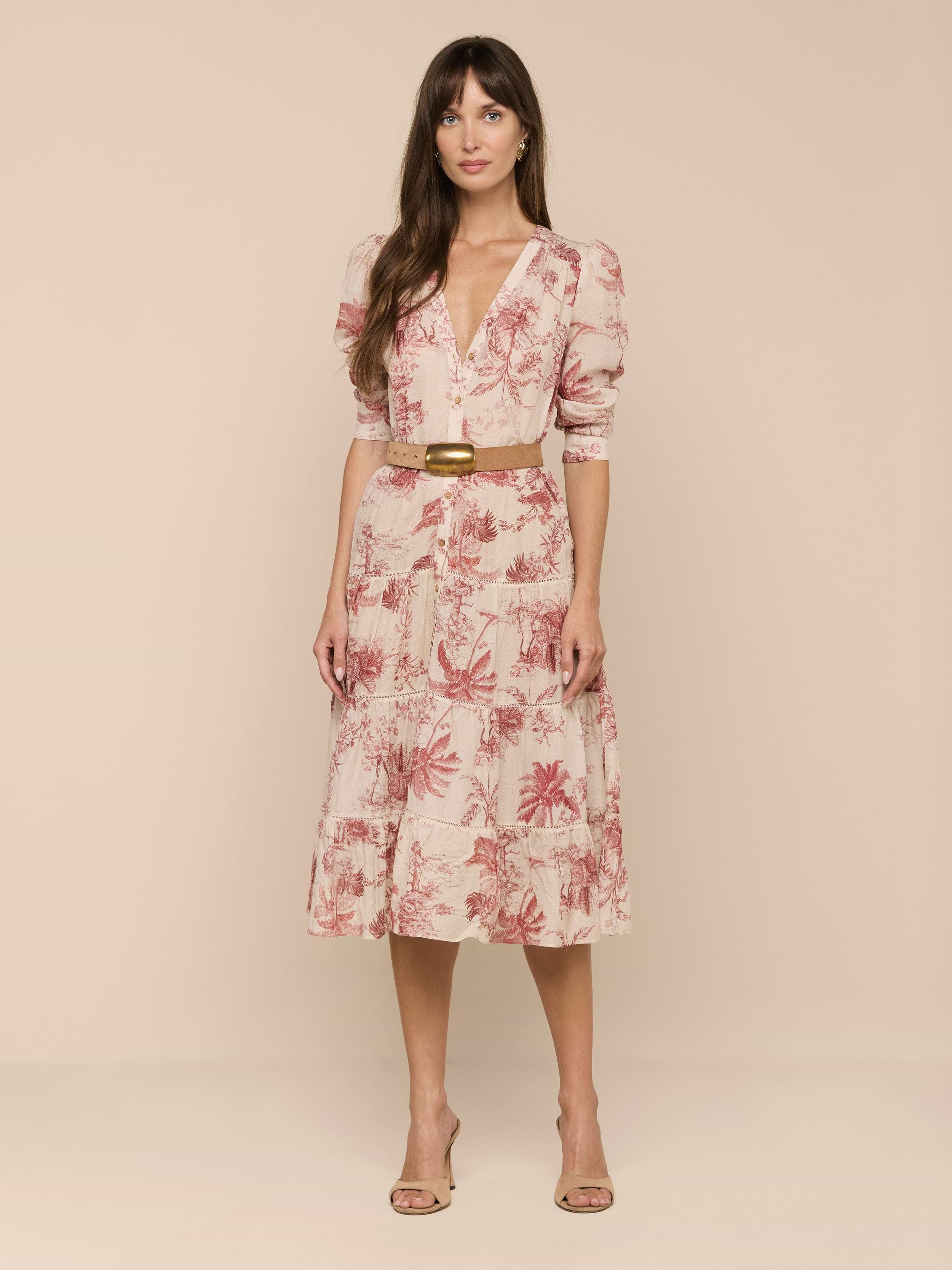 A woman with long brown hair stands against a plain white background wearing the Serafine Midi Dress, a light pink floral style featuring lightweight voile fabric, a V-neckline, puff sleeves, and a coordinating tan belt. She completes her look with nude heels.