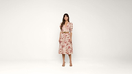 A woman stands against a plain white background, wearing a light pink, floral-patterned dress with puffed sleeves, a belt at the waist, and nude heels.