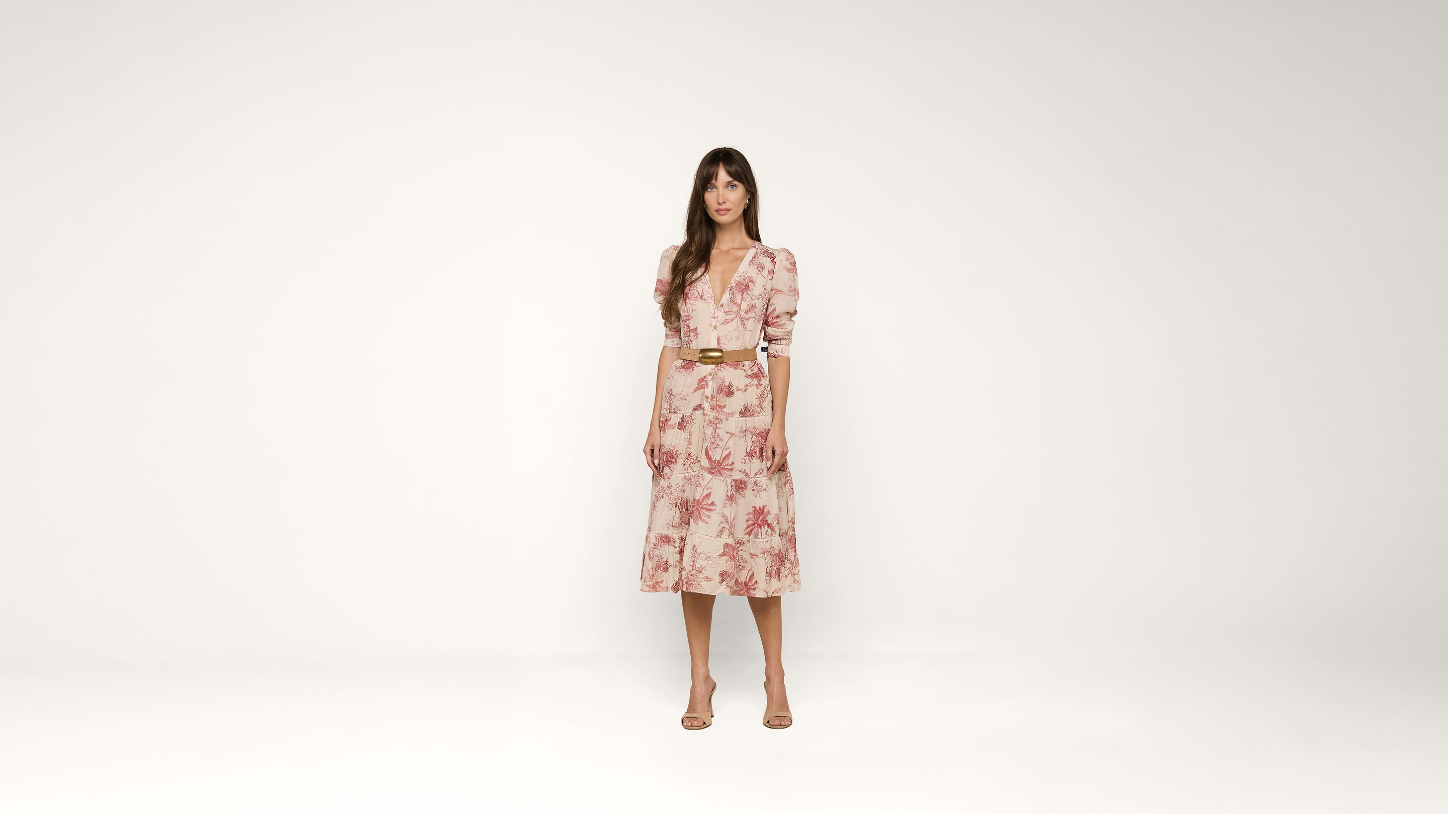A woman stands against a plain white background, wearing a light pink, floral-patterned dress with puffed sleeves, a belt at the waist, and nude heels.