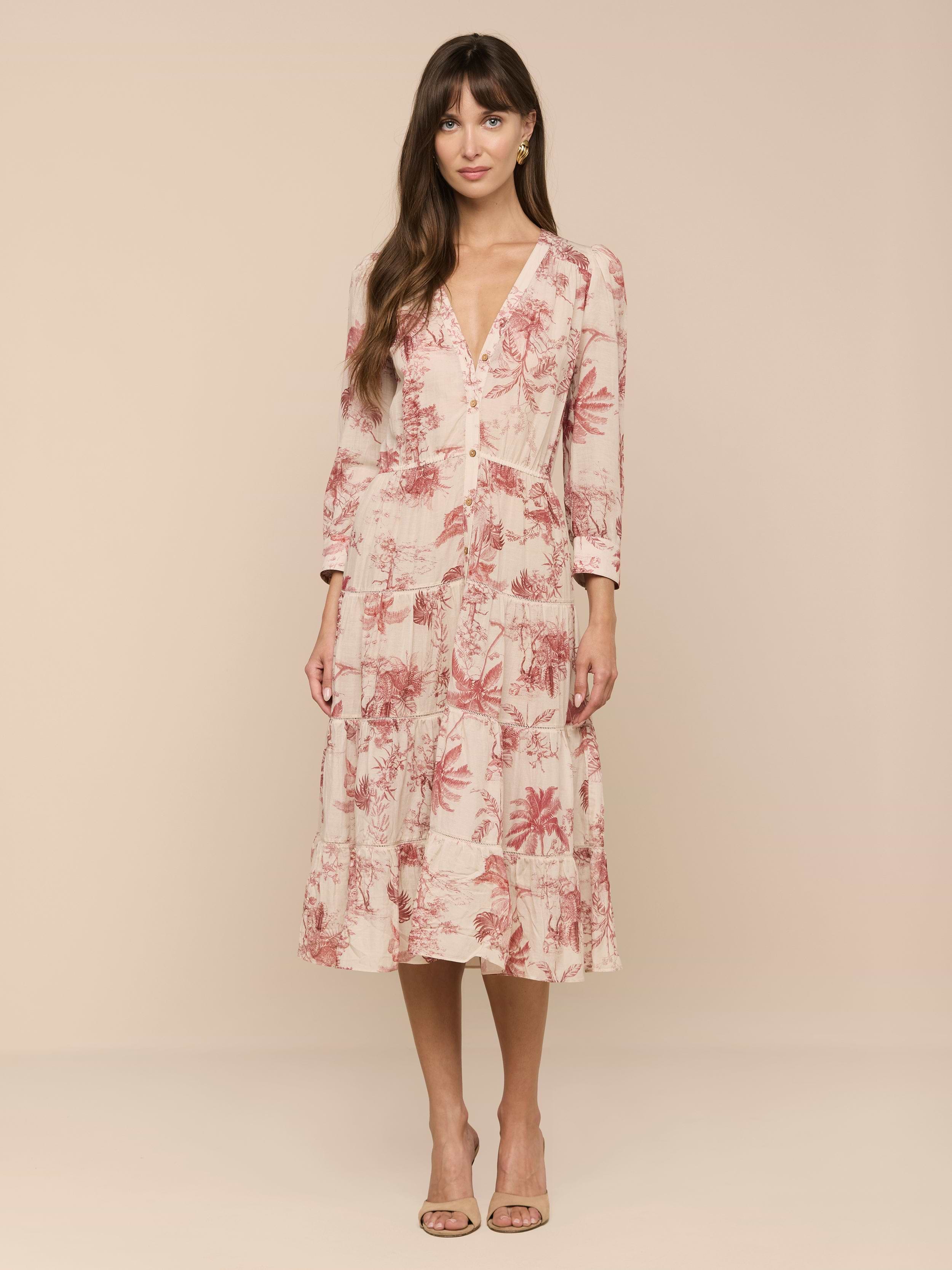 A woman stands against a plain white background wearing the Serafine Midi Dress—a light pink, long-sleeve midi dress with a red jungle print crafted from lightweight voile—paired with nude heels.