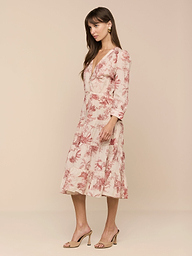 A woman stands in profile against a plain background, wearing the Serafine Midi Dress in light pink floral voile, paired with beige high-heeled sandals.