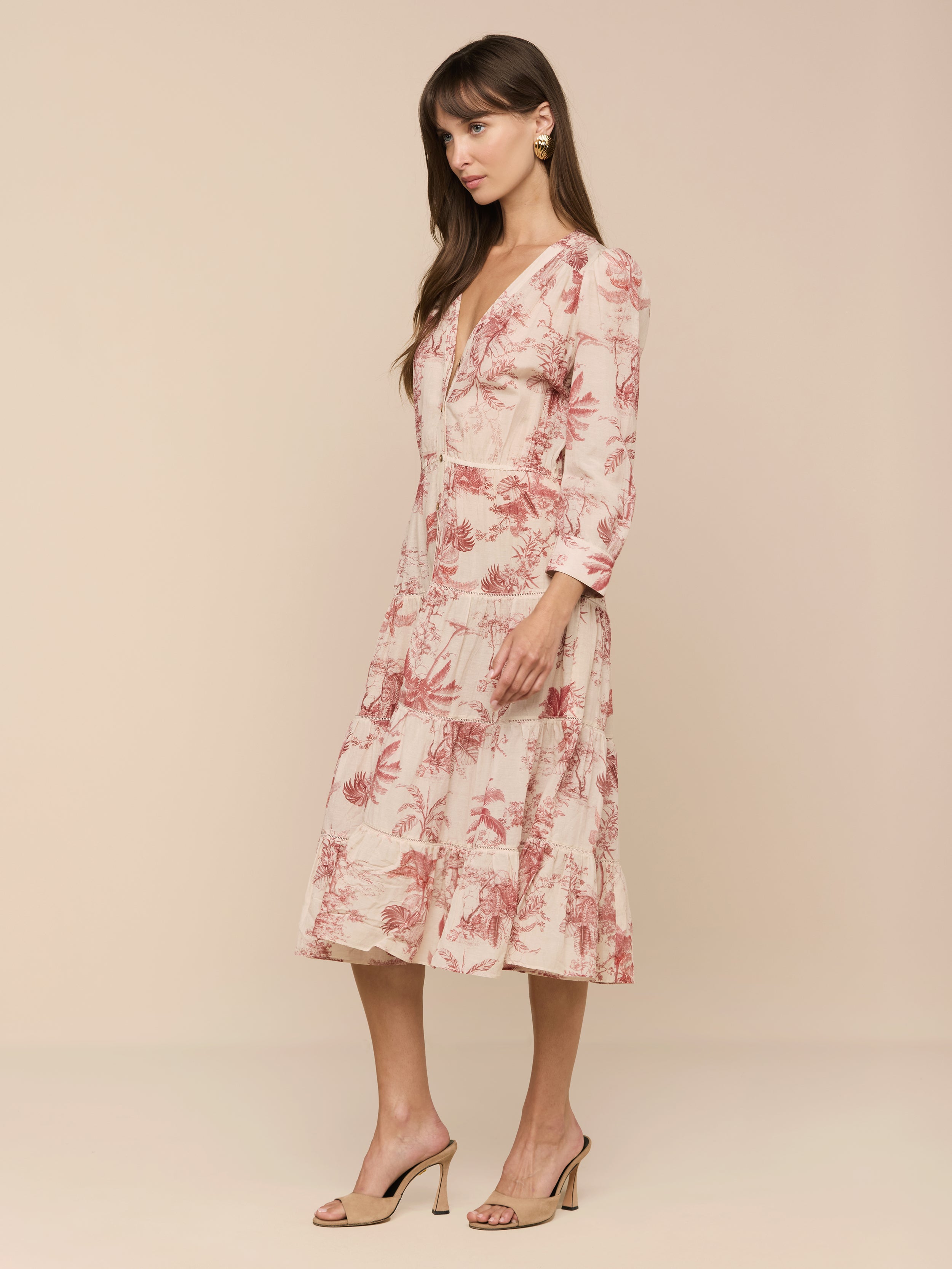 A woman stands in profile against a plain background, wearing the Serafine Midi Dress in light pink floral voile, paired with beige high-heeled sandals.