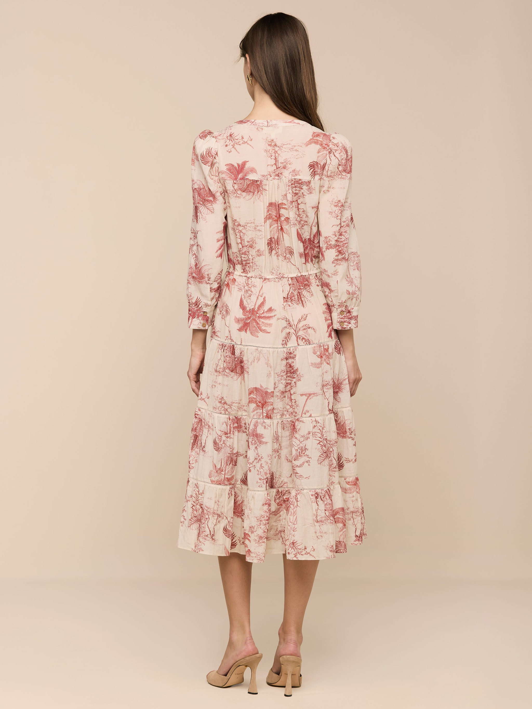 A woman stands with her back to the camera, wearing the Serafine Midi Dress—a light pink midi dress adorned with a red floral pattern—paired with beige heels. The dress is crafted from lightweight voile and photographed against a plain white background.