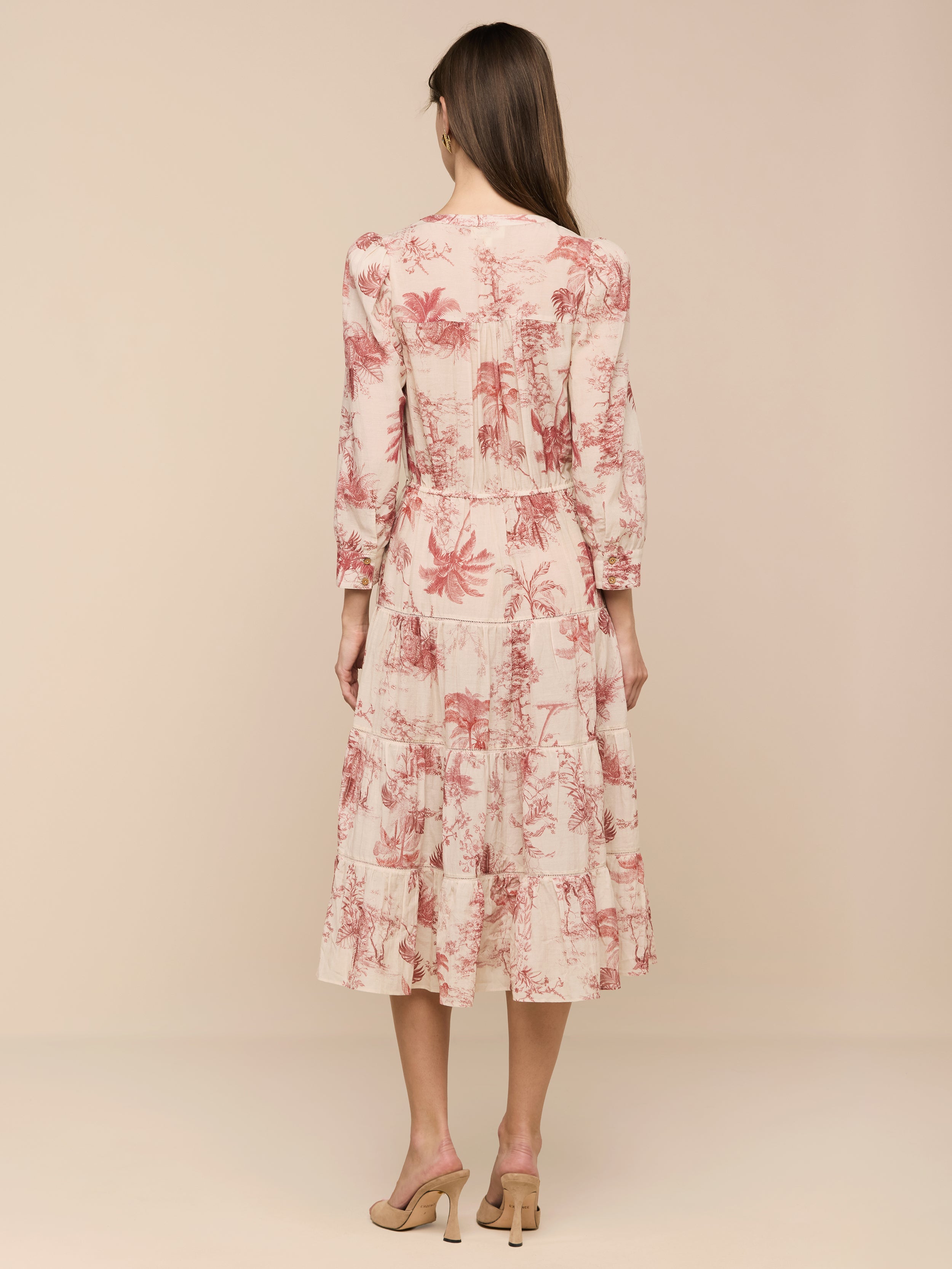 A woman stands with her back to the camera, wearing the Serafine Midi Dress—a light pink midi dress adorned with a red floral pattern—paired with beige heels. The dress is crafted from lightweight voile and photographed against a plain white background.