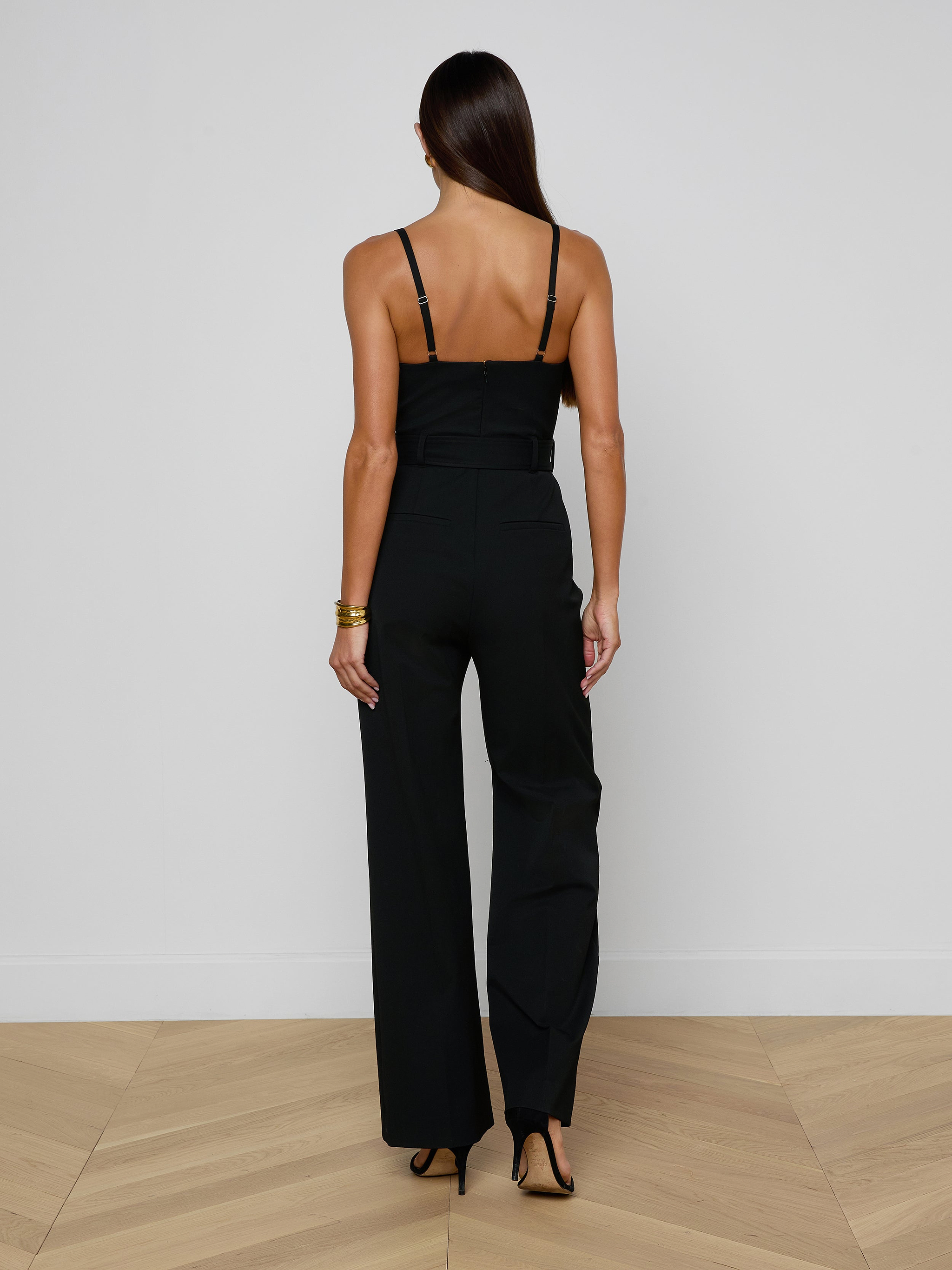 A woman stands with her back to the camera, wearing the Jessa Bustier Jumpsuit in black bi-stretch twill, paired with black heels, set against a plain light background.