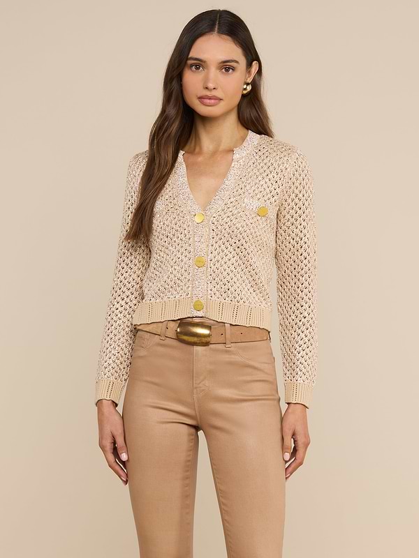 A woman stands facing forward, wearing the Blanca Sequin Cardigan—a neutral tan knit with gold button detailing—paired with tan high-waisted pants. Her long, straight brown hair and minimal jewelry highlight the look, while the plain light background emphasizes the cardigan as a perfect lightweight layer for everyday style.