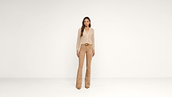 A woman stands against a plain white background wearing a light polka dot blouse, beige flared pants, and brown pointed shoes.