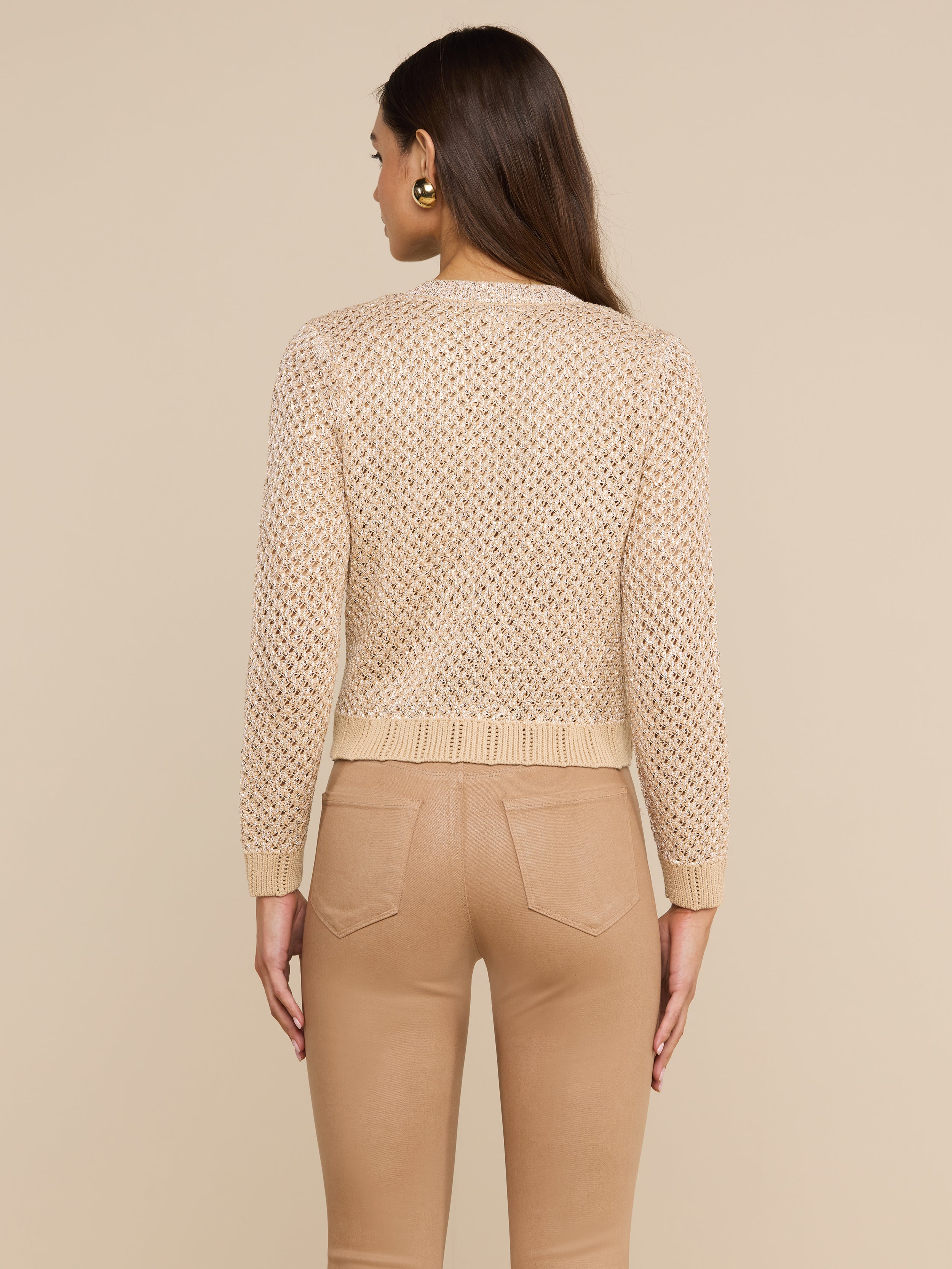 A woman with long brown hair stands facing away, wearing the Blanca Sequin Cardigan—a neutral tan sequined pointelle knit sweater—and tan pants with back pockets, against a plain light background.
