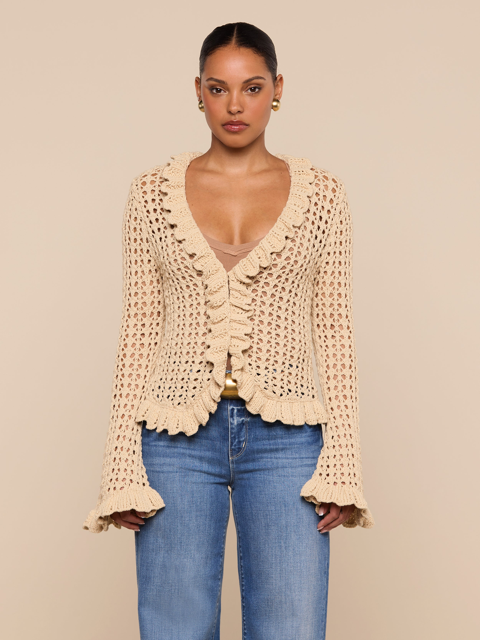 A woman is wearing the Solaris Crocheted Cardigan—a beige, ruffled, open-stitch crochet design with bell sleeves—paired with blue jeans and standing against a plain light background.