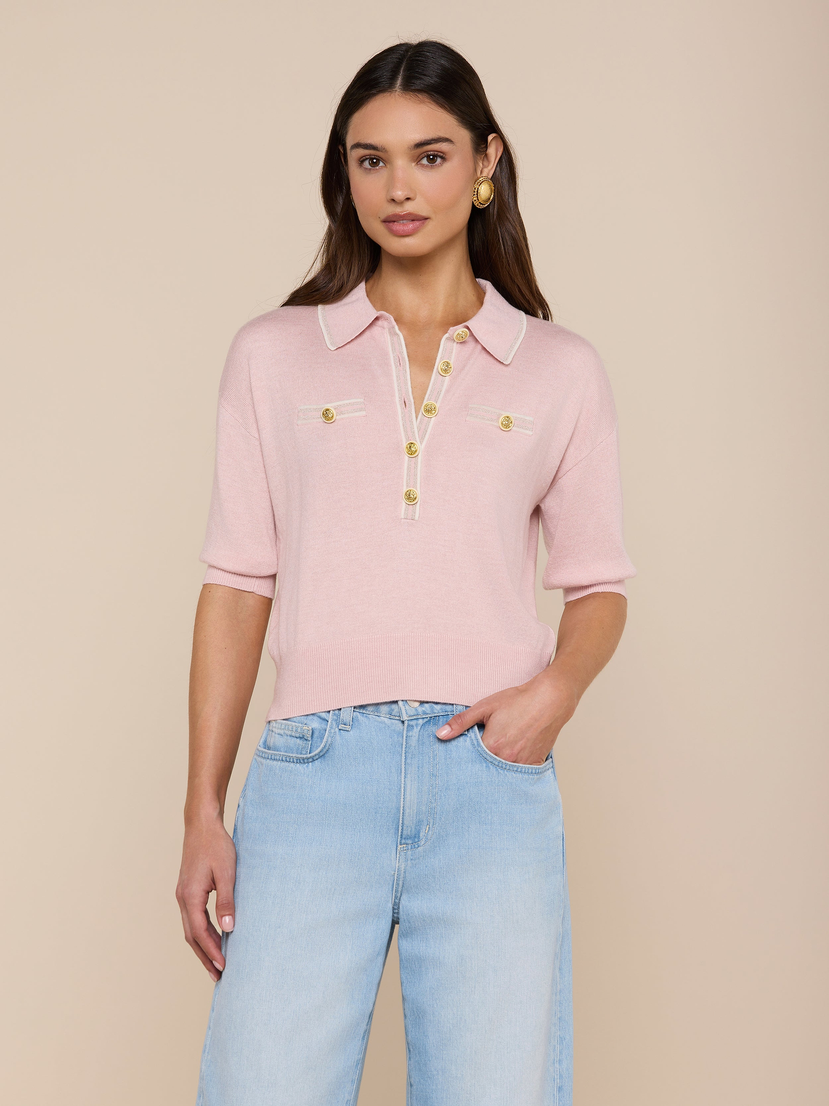 A woman stands against a plain white background wearing the Masaki Cropped Sweater—a light pink, featherweight knit polo top featuring gold buttons and front pockets—paired with light blue jeans.