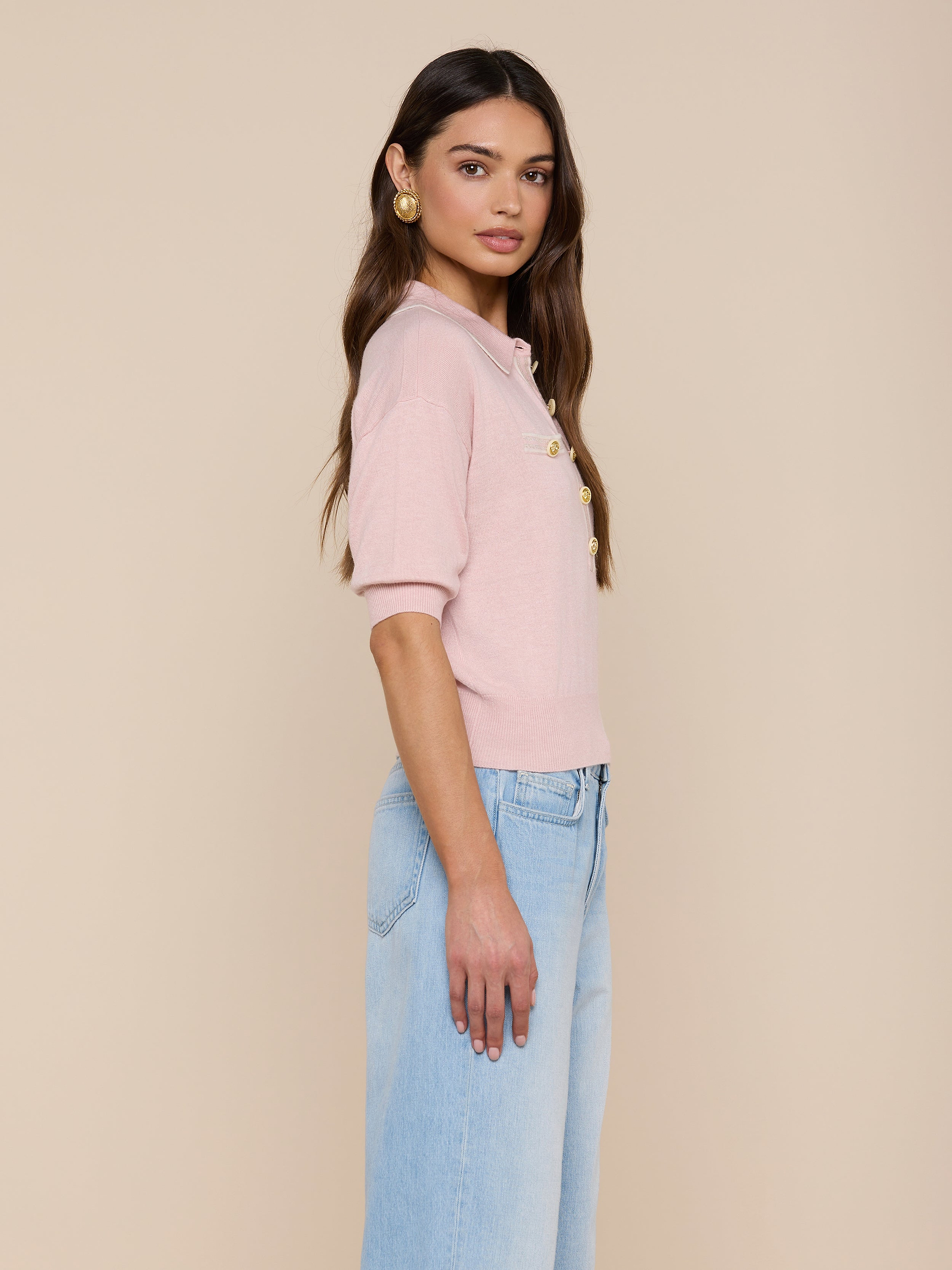 A woman with long, dark hair styled in loose waves stands in profile against a plain background, wearing the Masaki Cropped Sweater—a light pink featherweight knit polo top—paired with light blue high-waisted jeans and gold earrings.