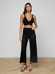 A woman stands against a plain white background wearing a black crop top, the Gaelle Pointelle Knit Pant in black featuring a sheer crochet knit and drawstring waist, and black high-heeled sandals.