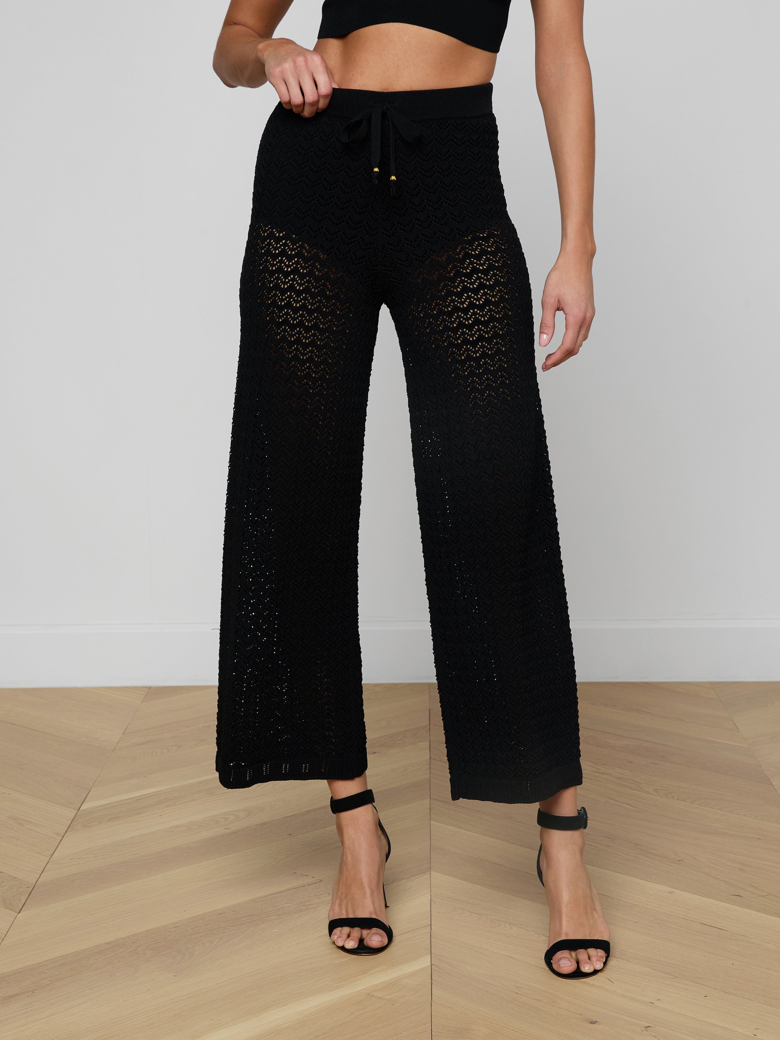 A woman wears the Gaelle Pointelle Knit Pant—a black, high-waisted, wide-leg knit pant with a drawstring waist—paired with a black crop top and black high-heeled sandals. Her face is not visible.