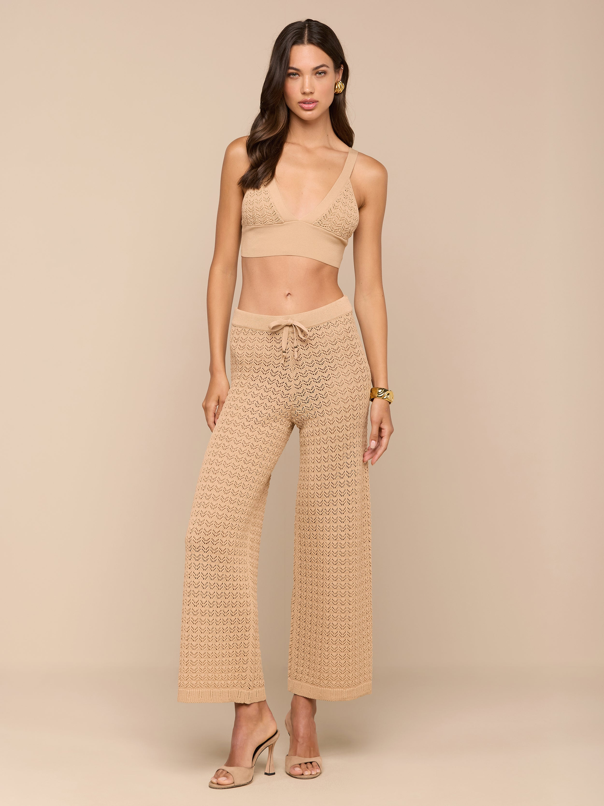Una donna su uno sfondo beige indossa una bralette alluncinetto abbinata e un completo Gaelle Pointelle Knit Pant con tacchi aperti. Ha i capelli mossi, orecchini e un braccialetto: perfetto per le vibrazioni delle vacanze.