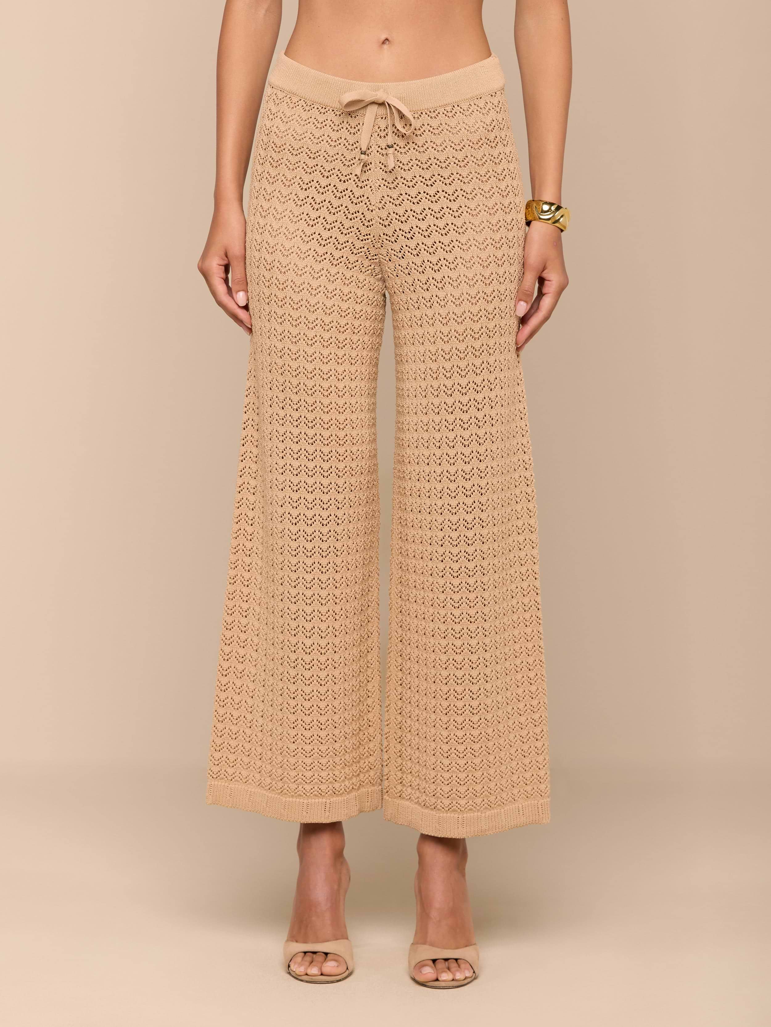 Una donna a piedi nudi modella il Gaelle Pointelle Knit Pant in tan, caratterizzato da una struttura a maglia alluncinetto, una silhouette a gamba larga e una coulisse in vita. Indossa un braccialetto doro; la parte superiore del corpo non è visibile. Questi pantaloni leggeri sono pronti per le vacanze.