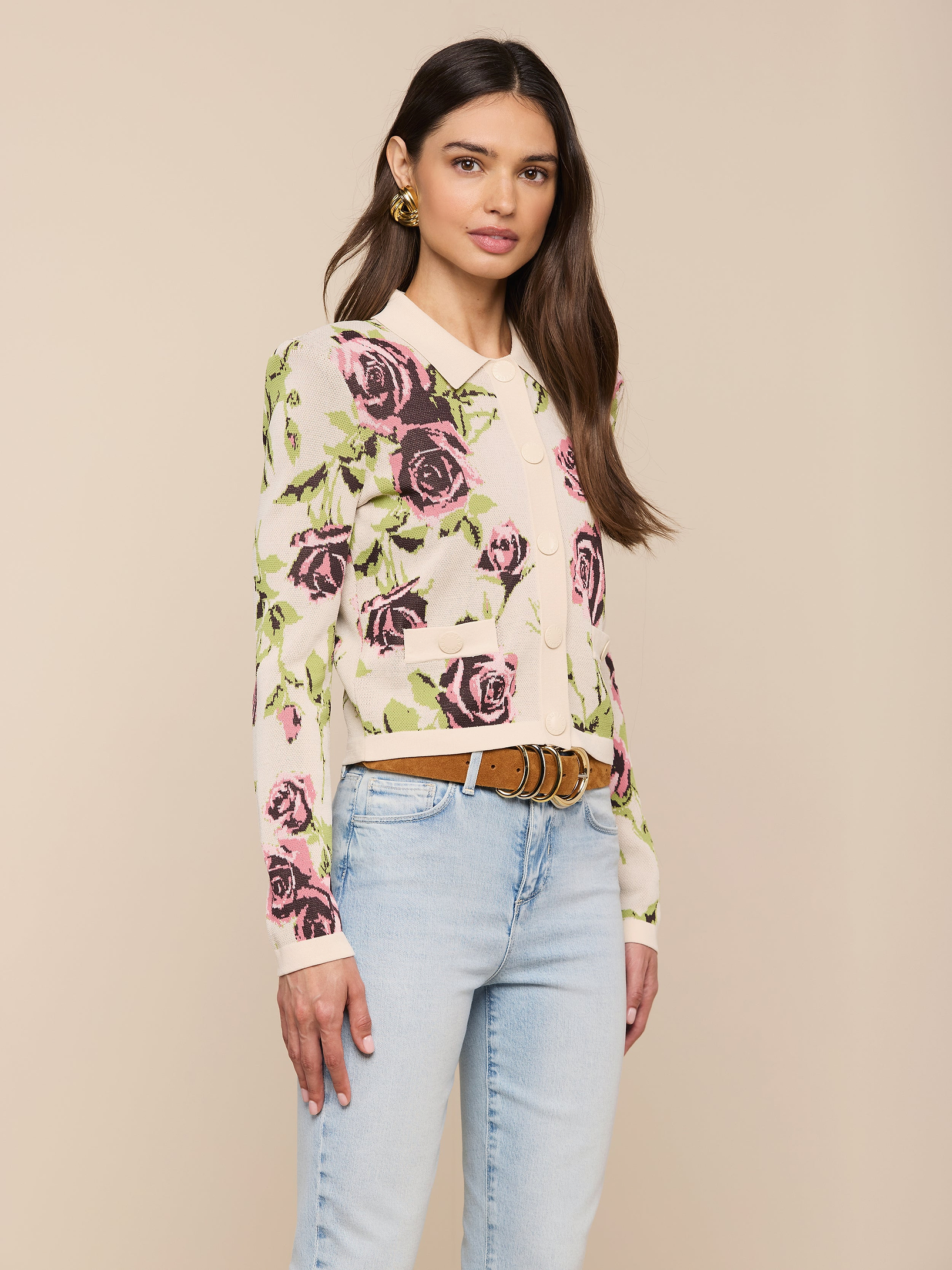 A woman stands against a plain light background, wearing the Osiris Jacquard Cardigan—a cream knit featuring a pink and purple rose motif—styled with light blue jeans, a brown belt, and gold earrings.