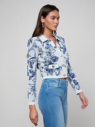A woman stands in profile against a plain light background, wearing the Osiris Jacquard Cardigan—a fitted blue and white floral-patterned knit button-up with silver buttons—paired with light blue high-waisted jeans.