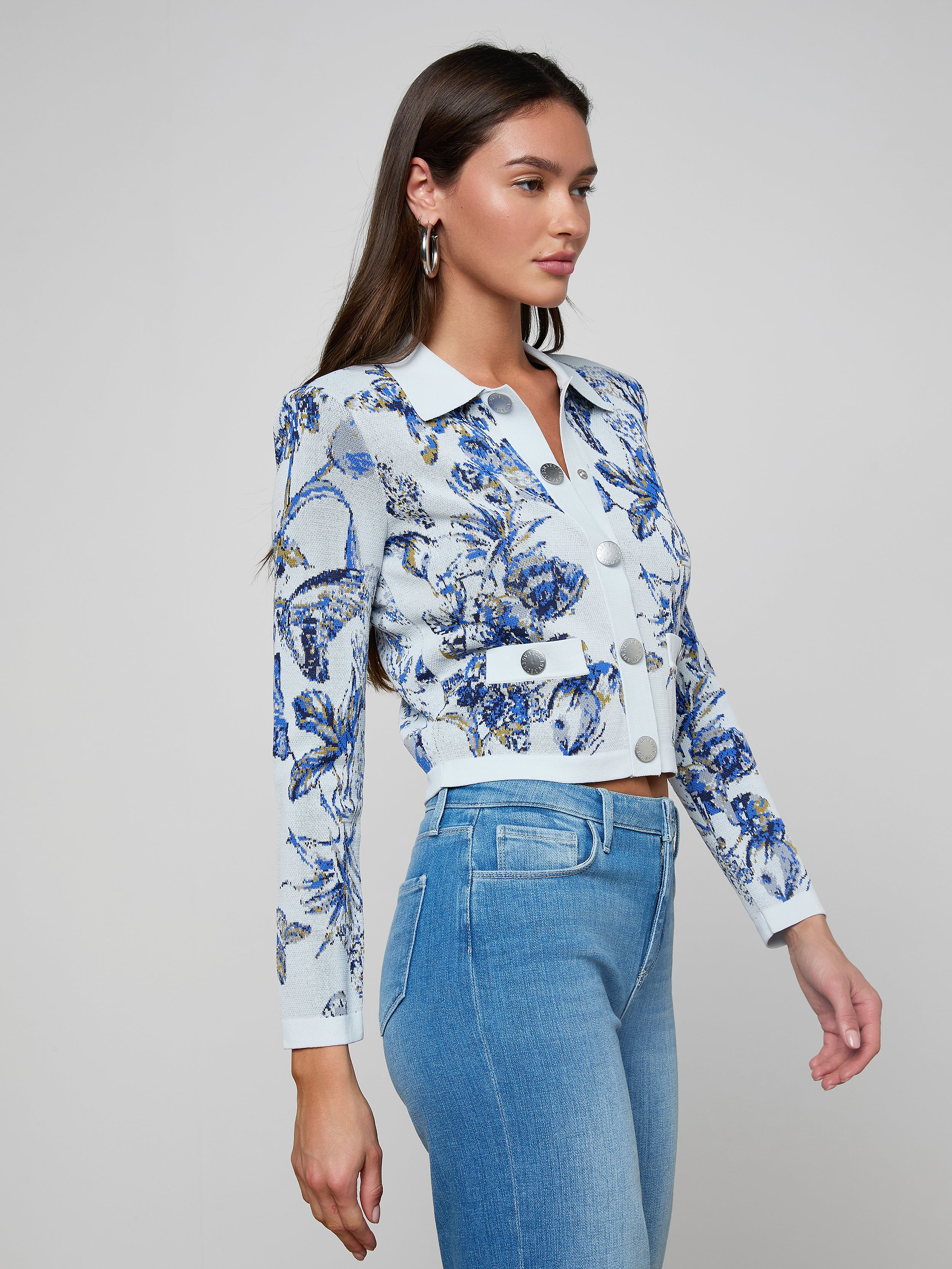 A woman stands in profile against a plain light background, wearing the Osiris Jacquard Cardigan—a fitted blue and white floral-patterned knit button-up with silver buttons—paired with light blue high-waisted jeans.