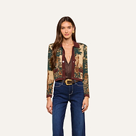 A woman with long brown hair wears the Osiris Jacquard Cardigan featuring a tapestry-inspired print with nature scenes, styled with a brown blouse, blue high-waisted jeans, and a belt with a gold buckle, standing against a plain off-white background.